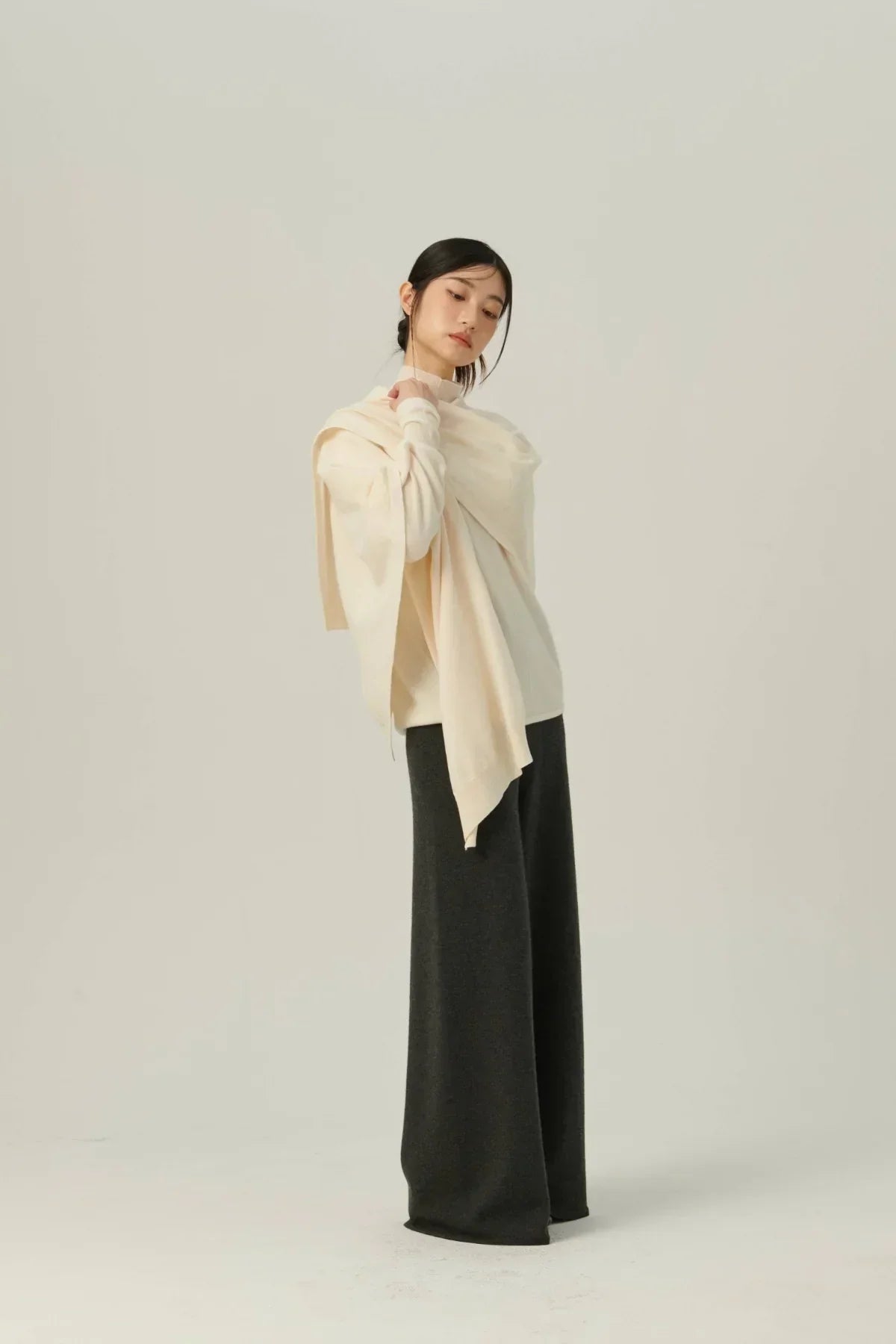 Woman wearing ivory multiway knit cape and wide-leg dark trousers standing against neutral background