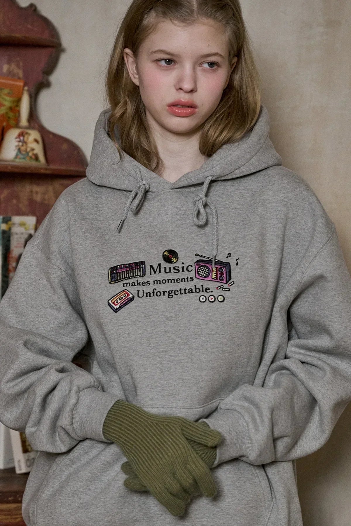Young woman wearing gray hoodie with music-themed embroidery and green knit gloves, indoor setting