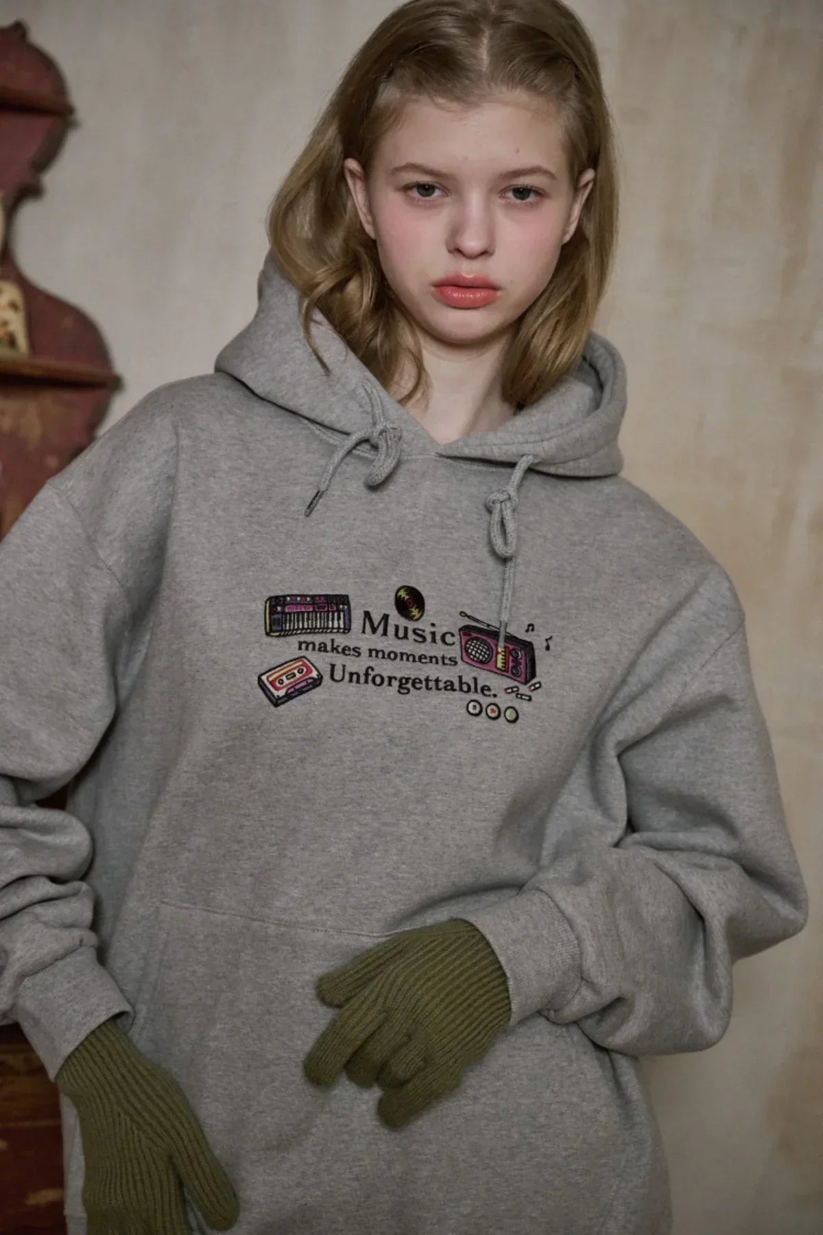 Young woman wearing gray hoodie with music-themed embroidered text and green gloves indoors