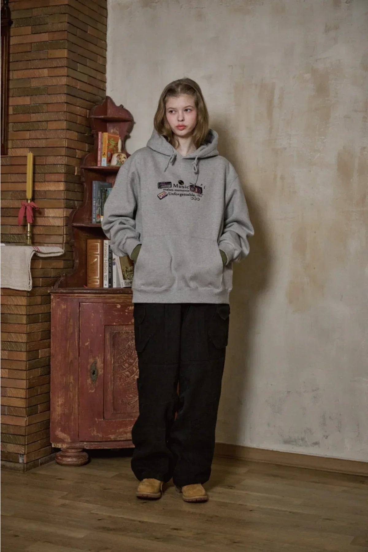young woman wearing gray embroidered music hoodie and black cargo pants standing indoors by vintage cabinet