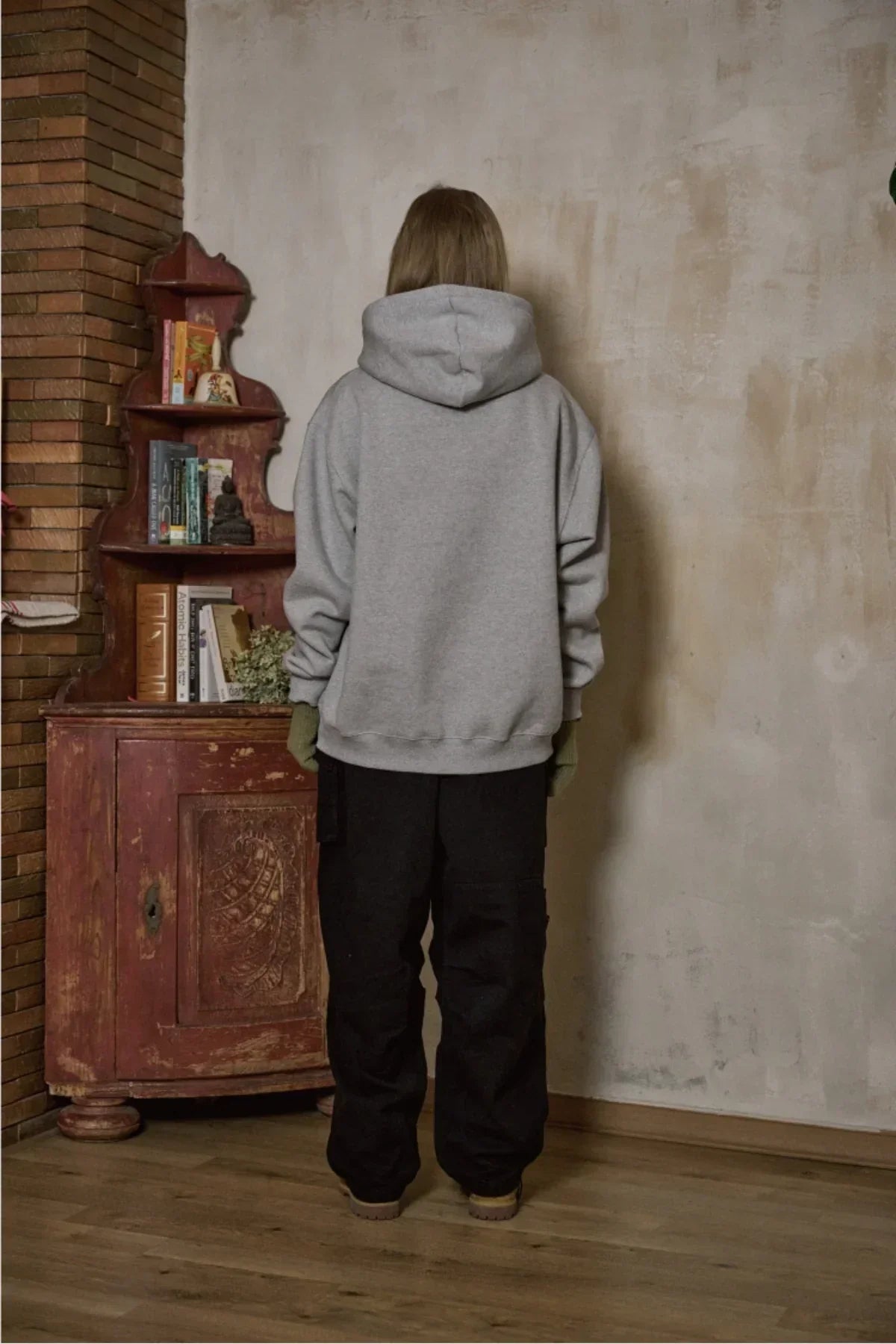 Person wearing gray hoodie and black pants standing indoors near vintage wooden cabinet