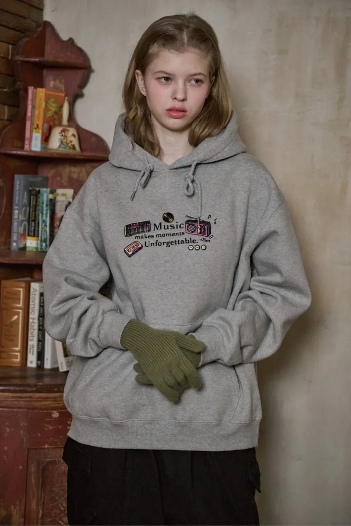 Young woman wearing gray embroidered hoodie with music-themed design and green gloves indoors
