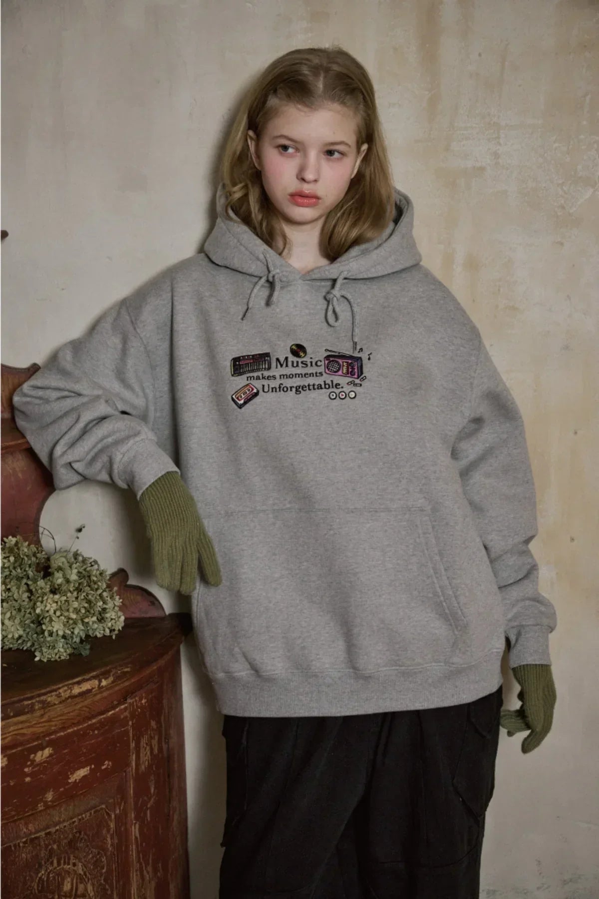 Young woman in gray music-themed hoodie with green gloves posing indoors by rustic wooden table