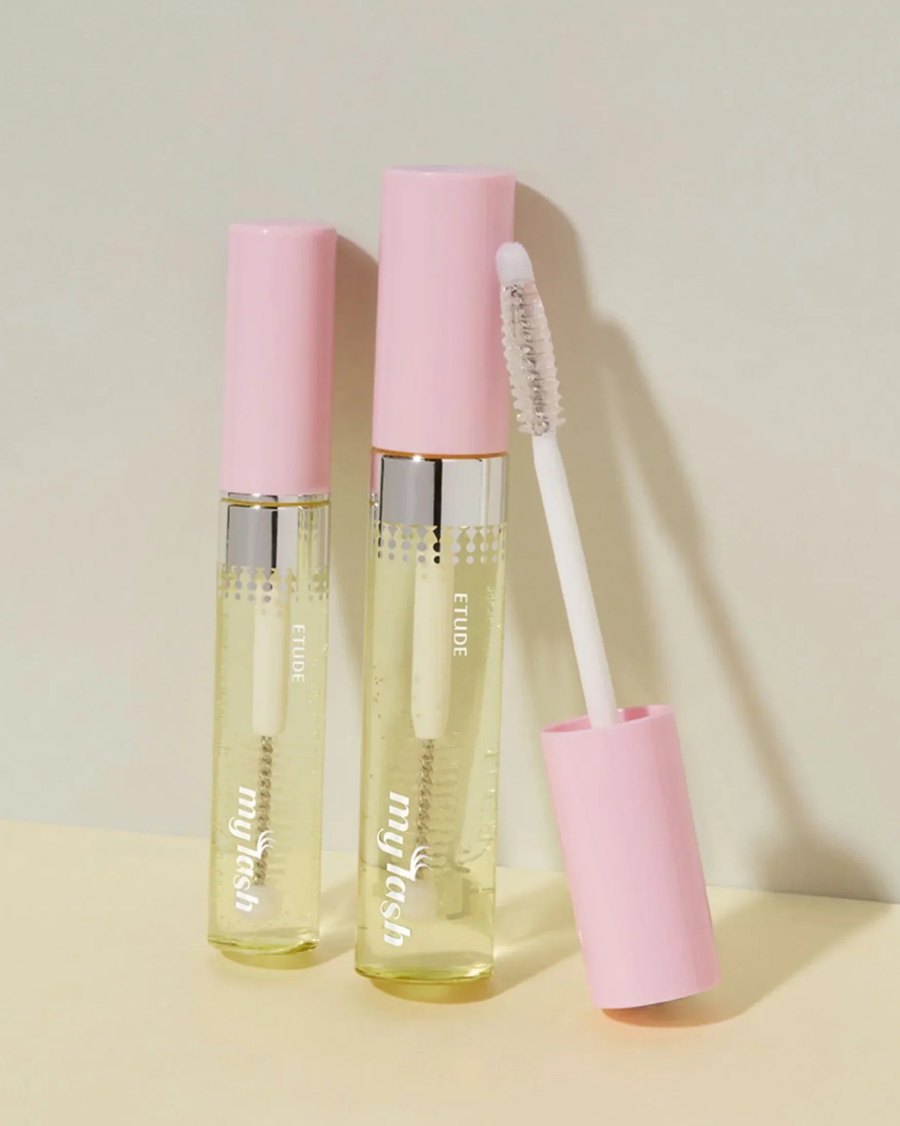 Two clear eyelash serum tubes with pink caps, one open showing the brush applicator on beige background