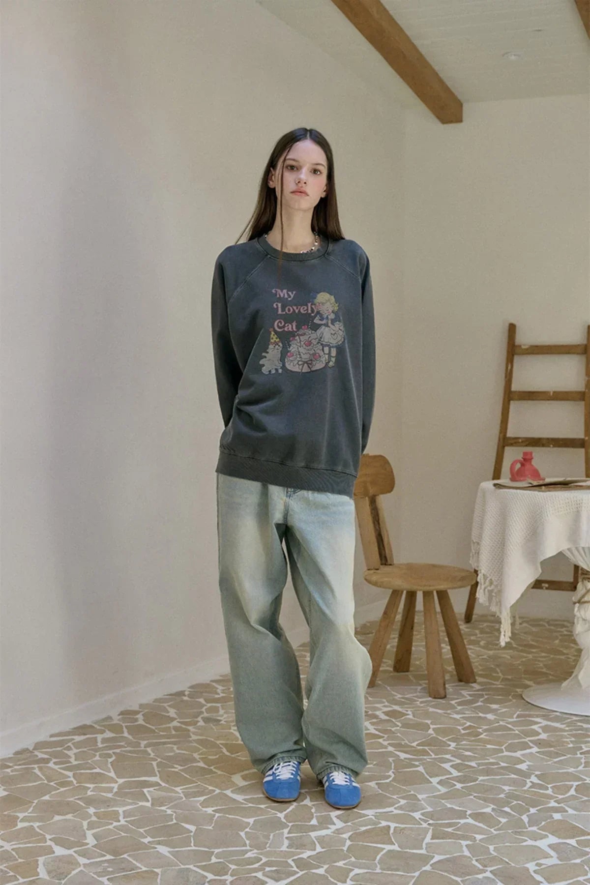 Woman wearing charcoal raglan sweatshirt with cartoon cat and text My Lovely Cat, loose light blue jeans, and blue sneakers in a minimal room