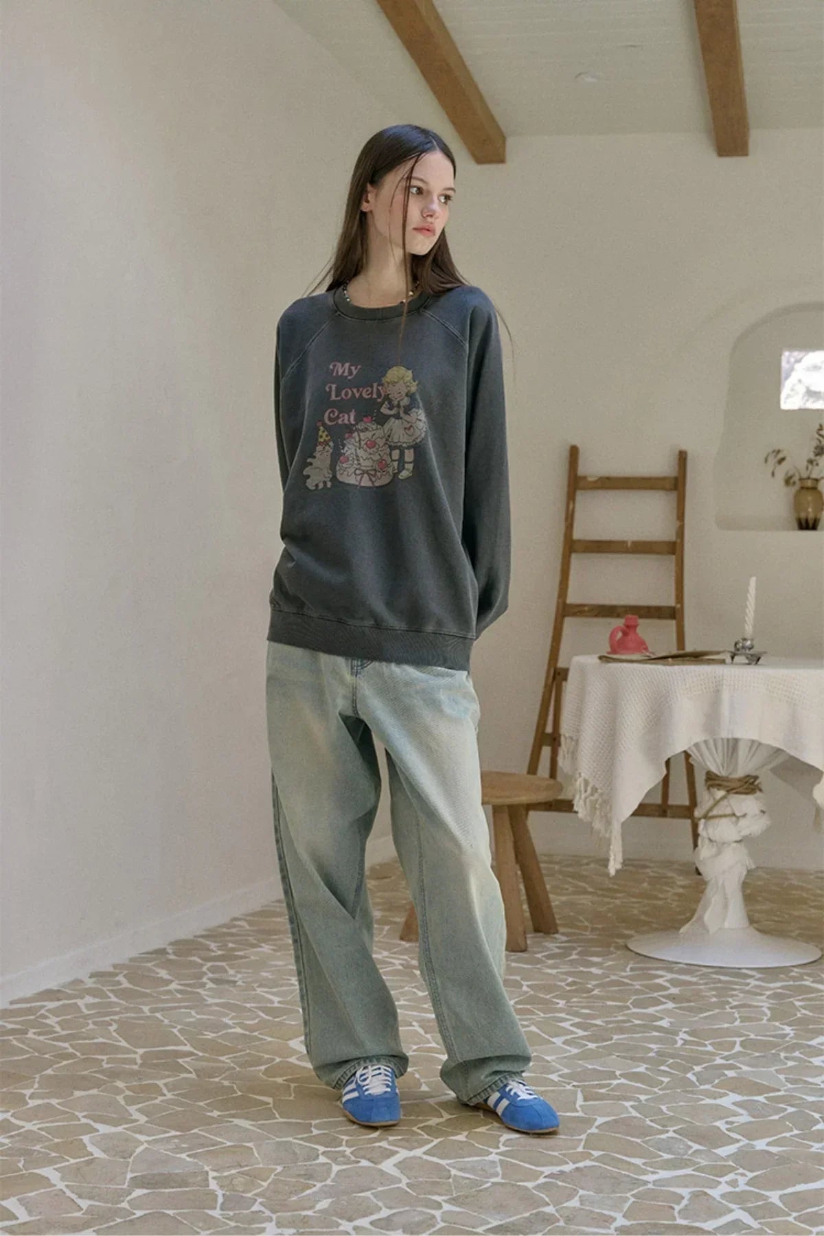 Young woman wearing dark gray sweatshirt with "My Lovely Cat" print, light blue jeans, and blue sneakers in a cozy room