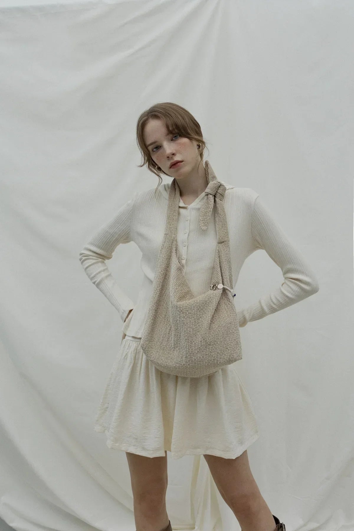 Young woman wearing beige knitted natural knot bag over white ribbed cardigan and skirt against white backdrop