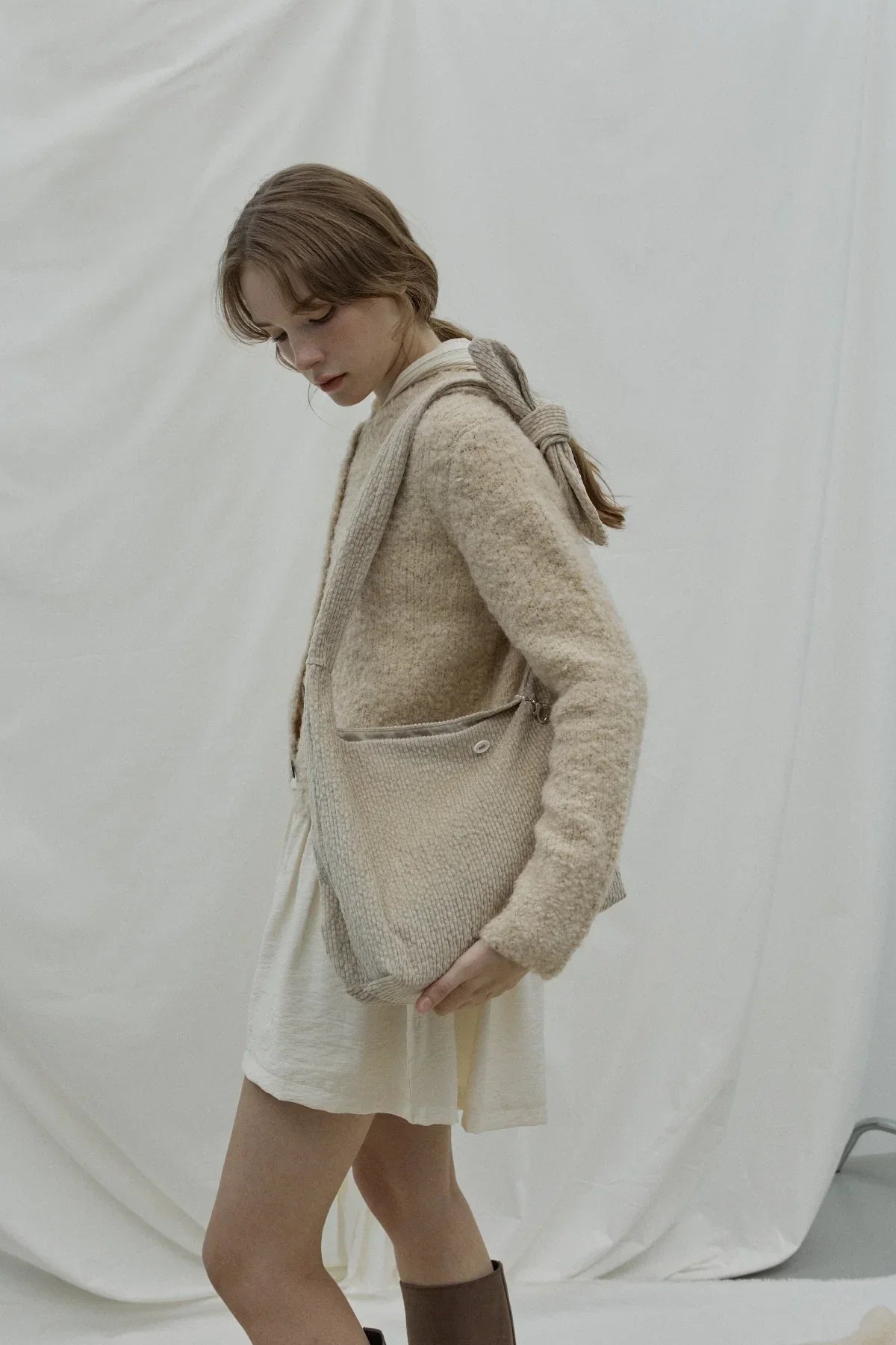 Woman in beige fuzzy cardigan and white dress carrying a beige knit knot shoulder bag in studio