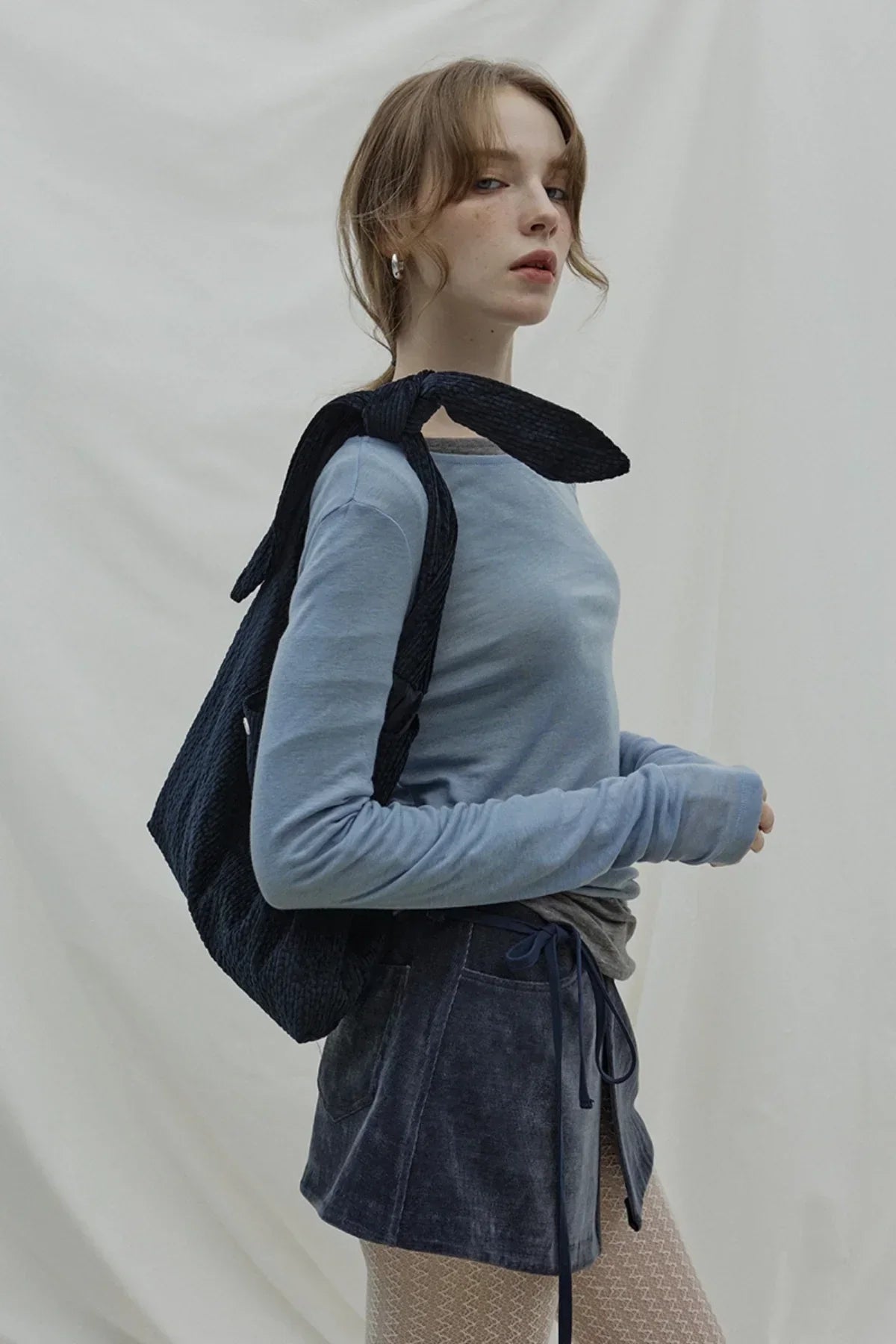 Woman wearing light blue long sleeve top and dark navy natural knot bag over shoulder against plain background
