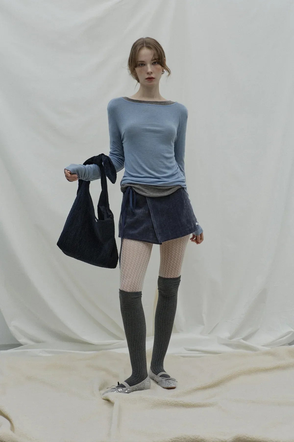 Woman in blue long-sleeve top, navy wrap skirt, gray knee-high socks, and leopard print flats holding a dark navy knot bag