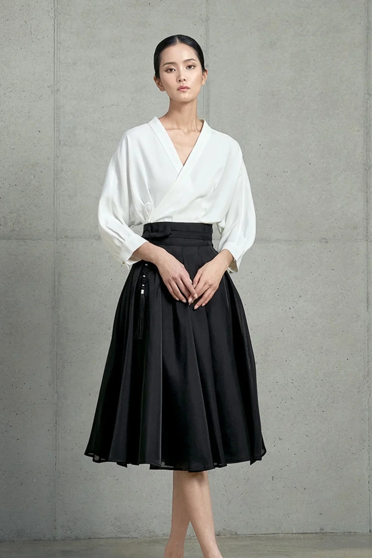 Model wearing black pleated mid-length skirt with tassel detail and white wrap blouse against concrete wall