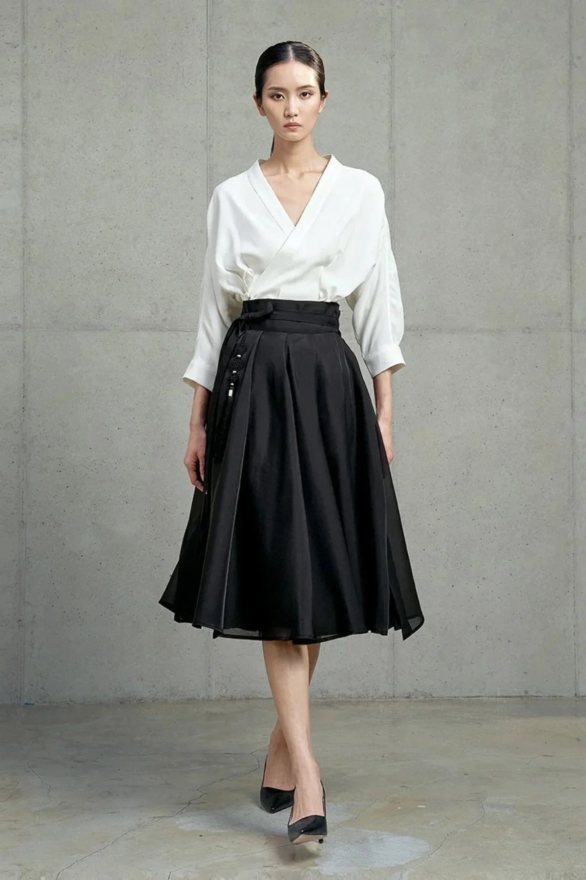 Model wearing black midi skirt with high waist and white blouse, posing against gray concrete wall