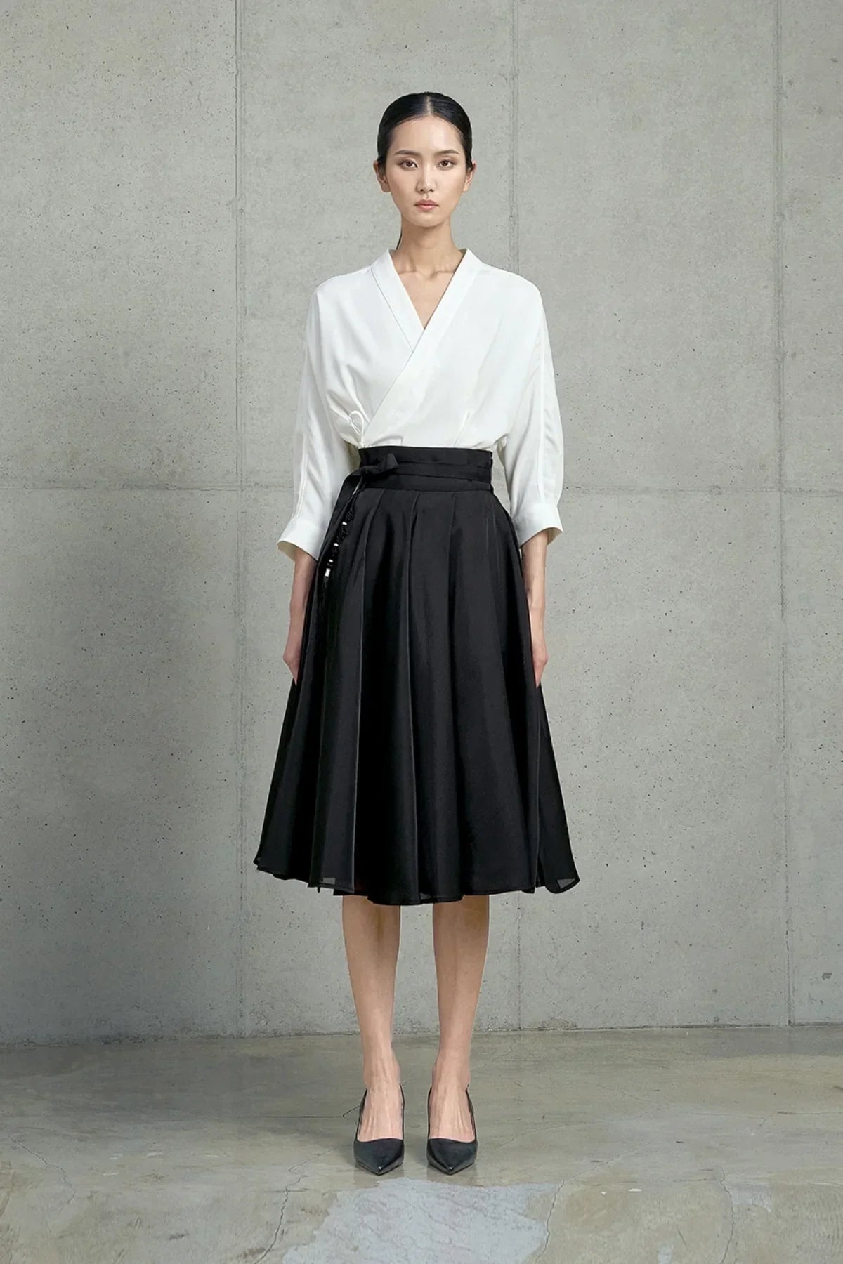 Model wearing black pleated midi skirt and white V-neck blouse against gray concrete wall