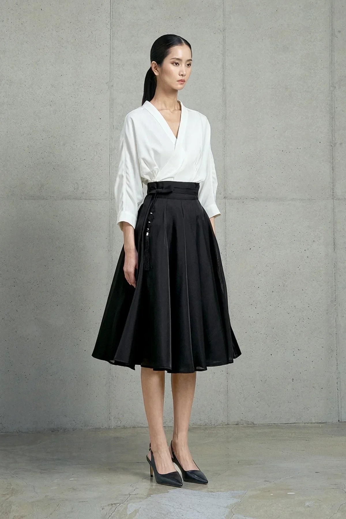Model wearing a black midi skirt with pleats and tassel detail paired with a white wrap blouse and black heels