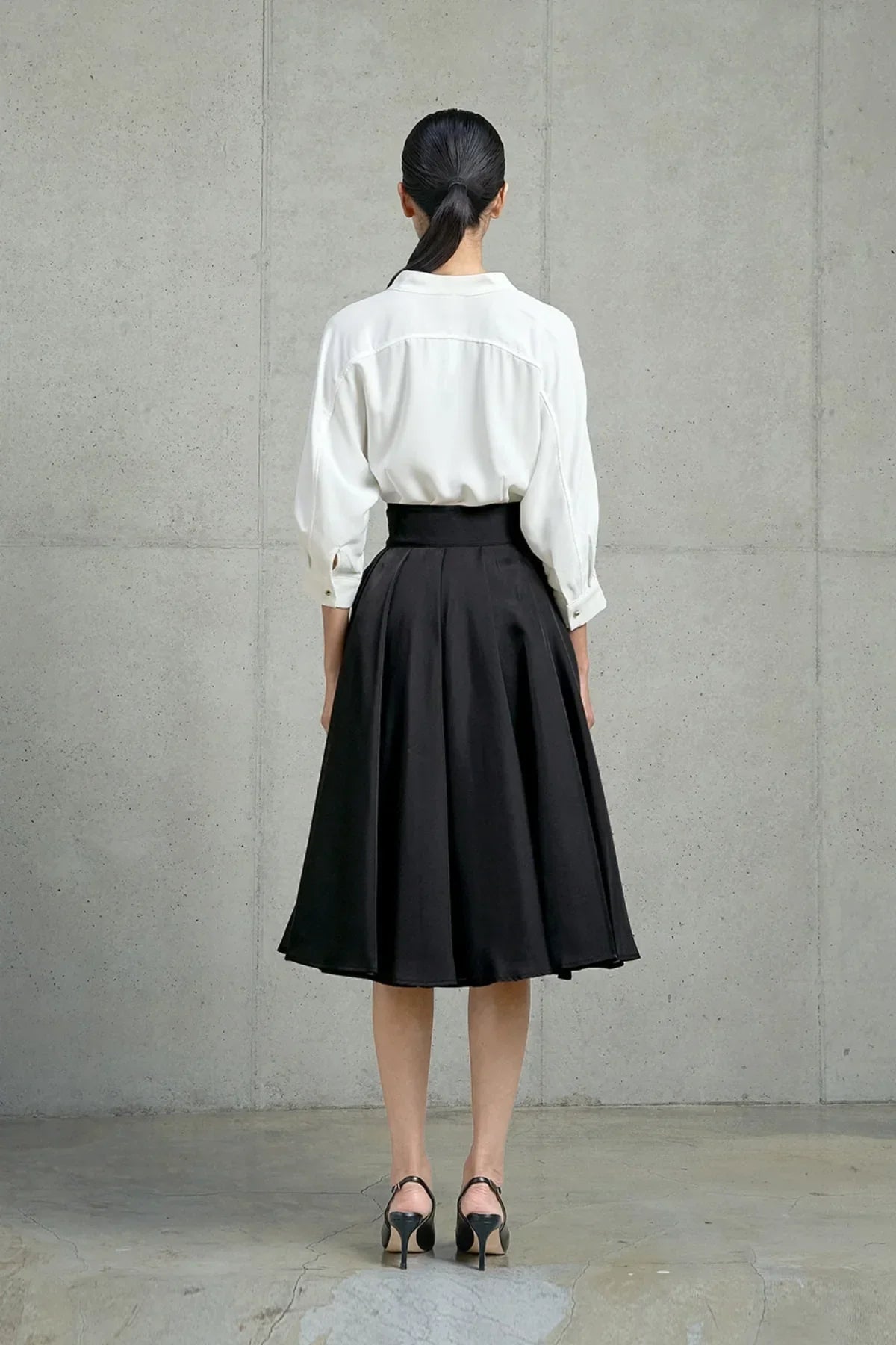 Rear view of woman wearing black pleated midi skirt, white blouse, and black heels against concrete wall