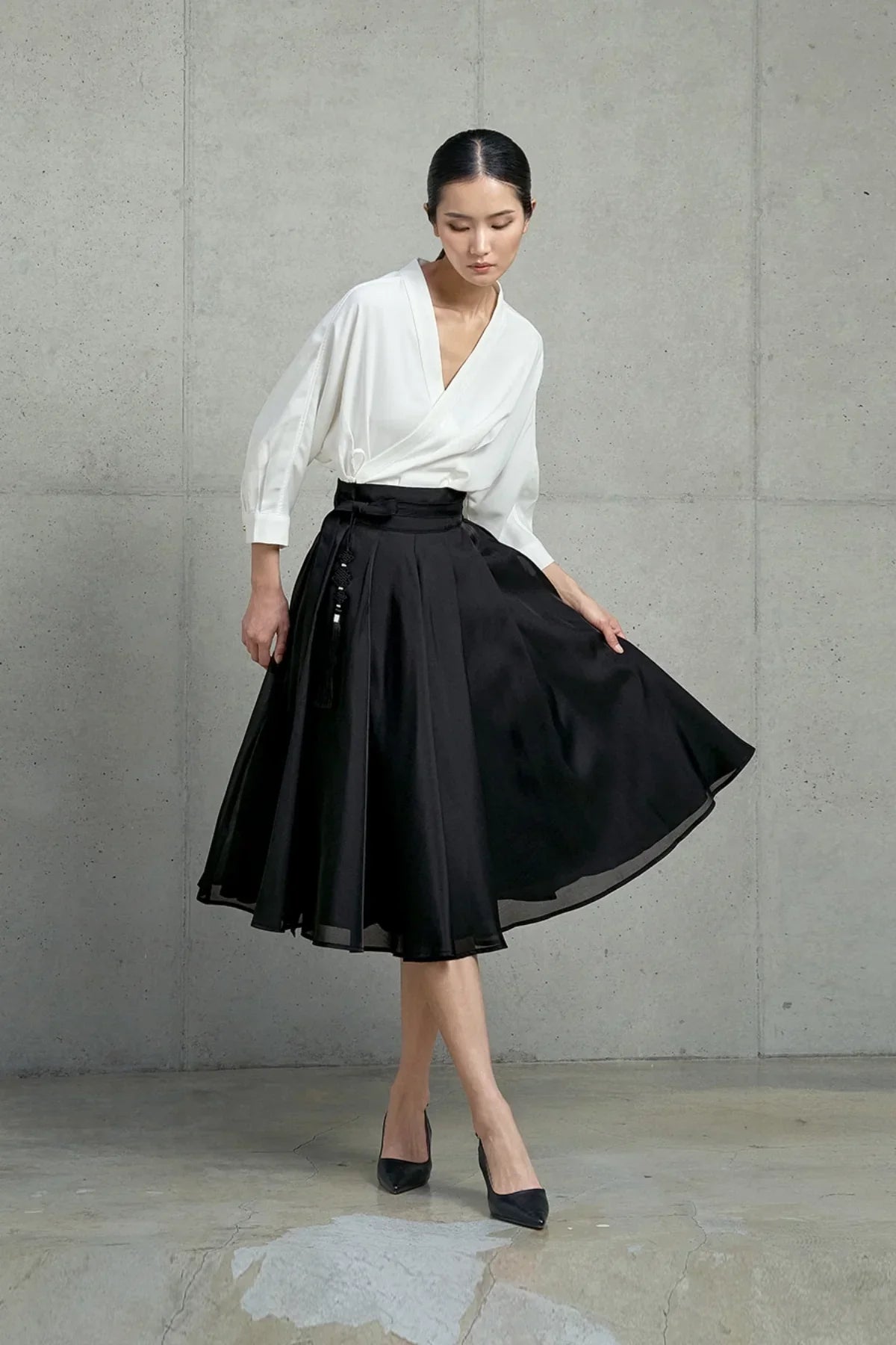 Woman wearing a white wrap blouse and a high-waisted black midi skirt with tassel detail, posing in front of a concrete wall