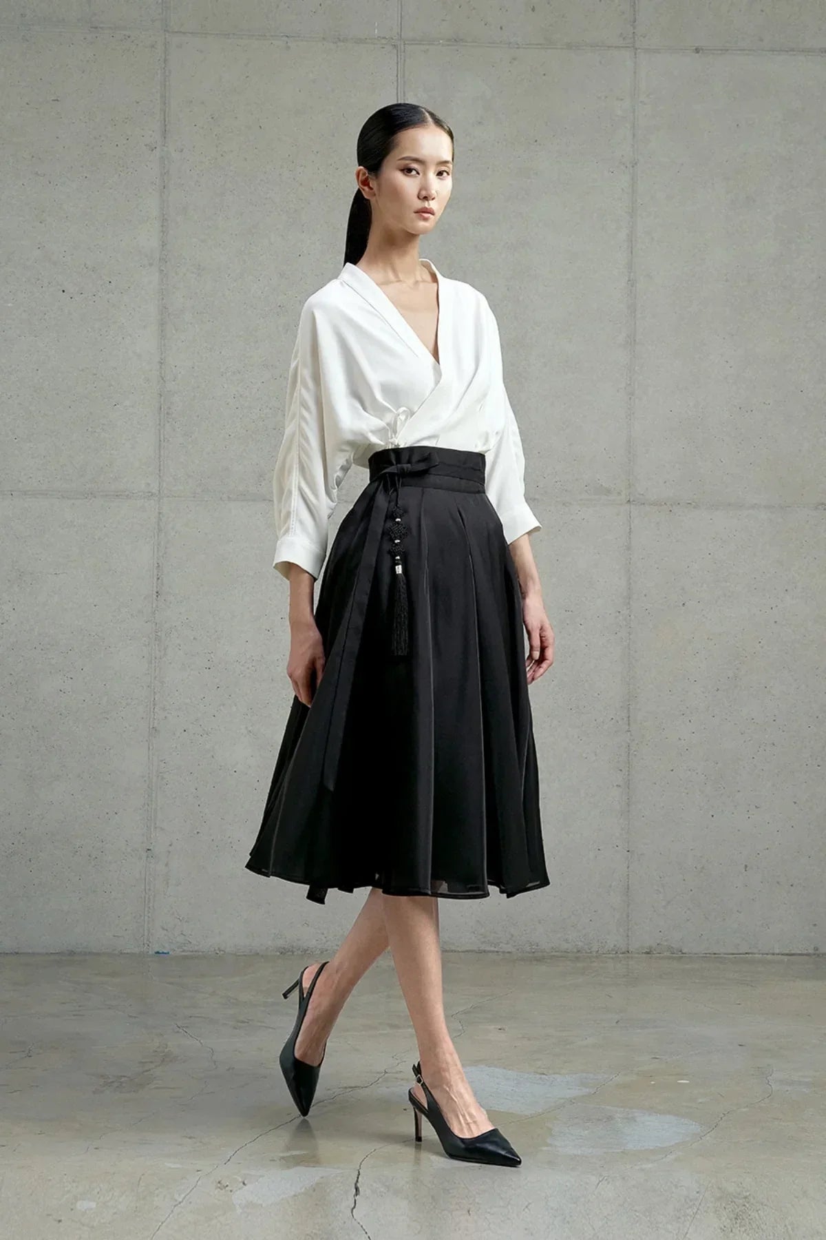 Model wearing black pleated skirt with tassel detail and white wrap blouse in minimalist studio