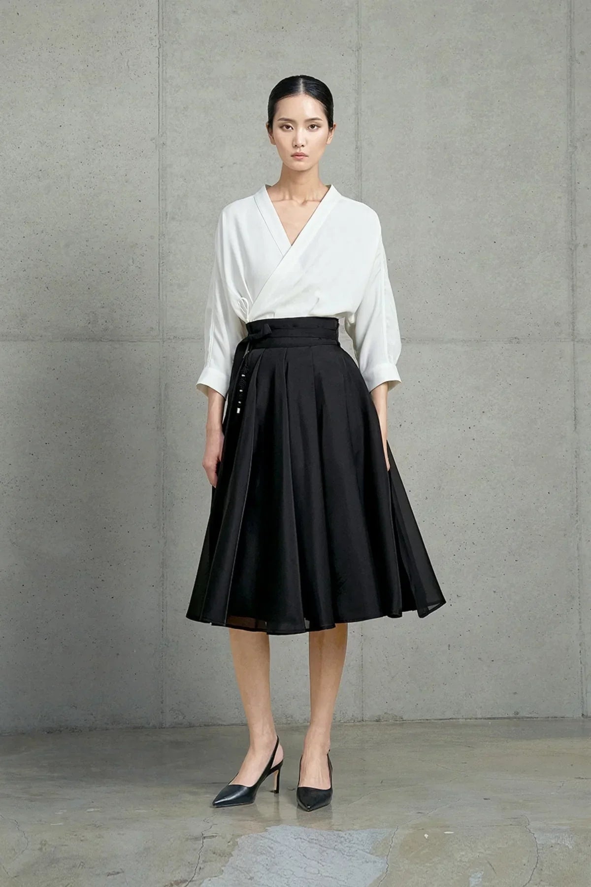 Model wearing a white wrap blouse with a high-waisted black A-line midi skirt and black heels in a minimalist studio