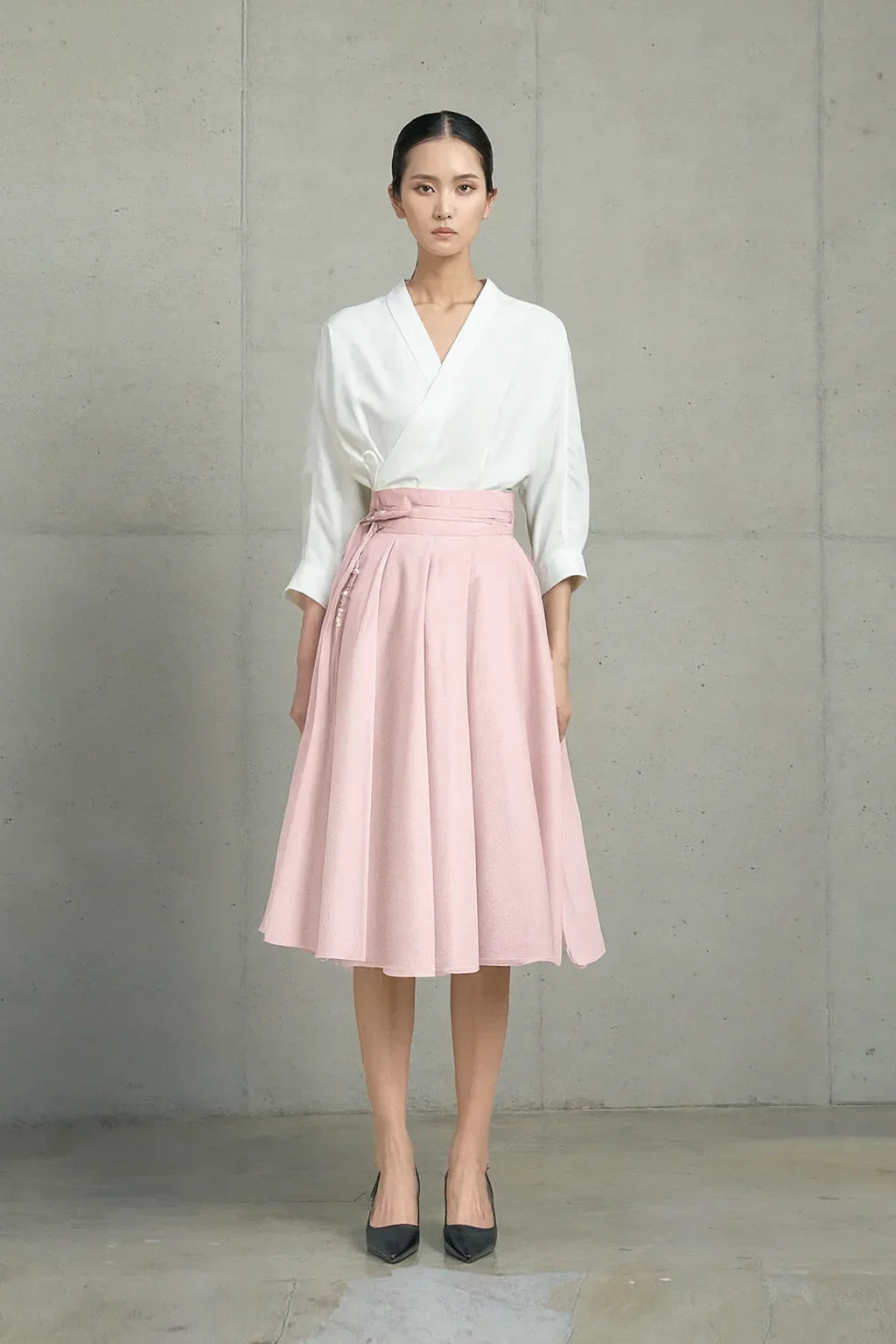 Model wearing blush pink midi skirt with pleats and high waist belt, paired with white wrap top and black heels