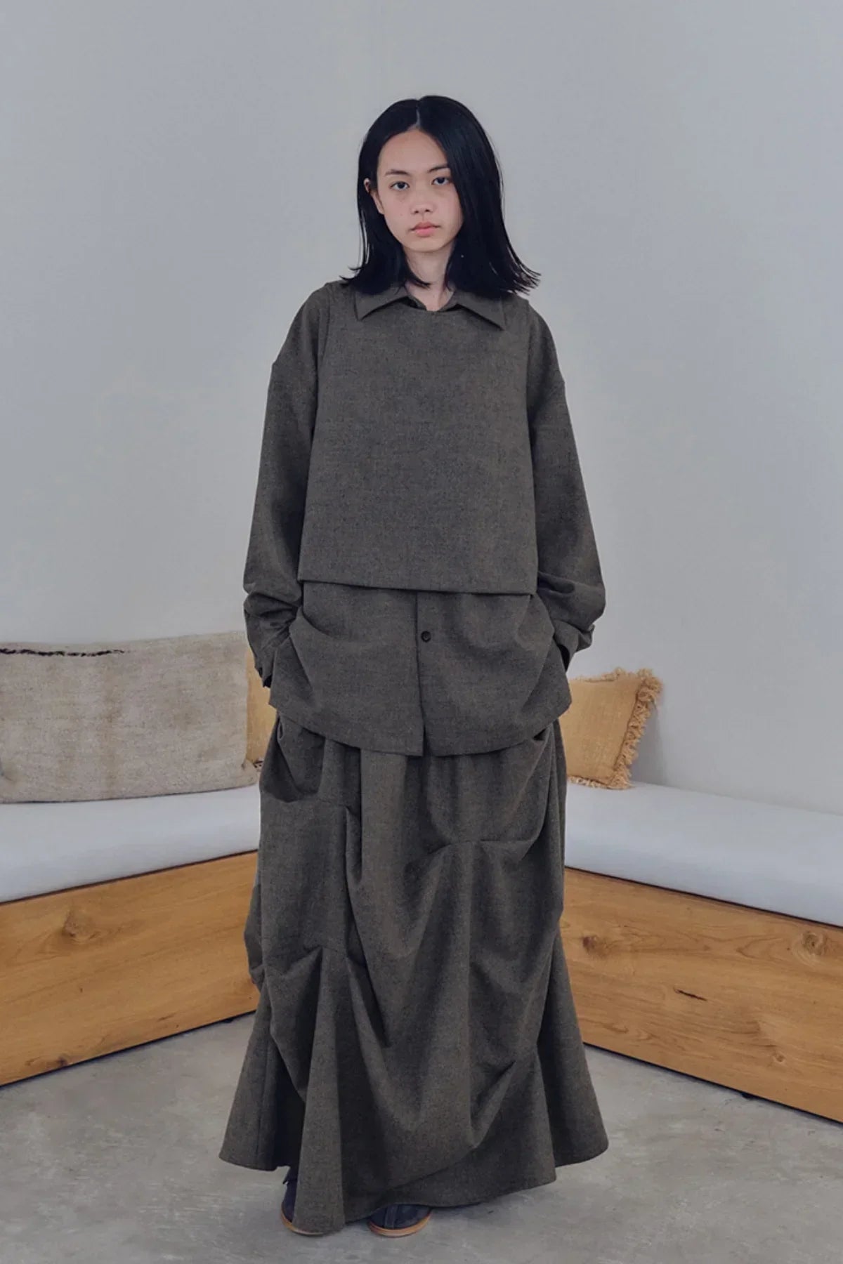 Woman wearing dark brown oversized long skirt and matching loose jacket indoors with wooden bench and cushions