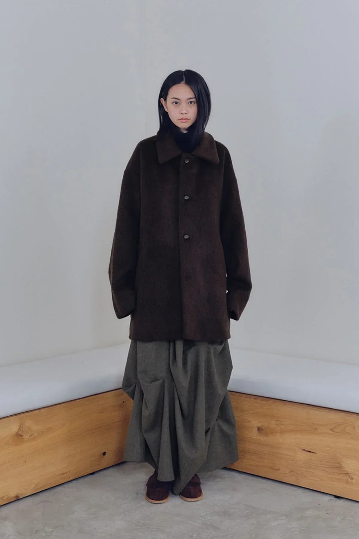 Model wearing dark brown oversized coat and olive draped skirt standing in minimalist corner with wooden bench