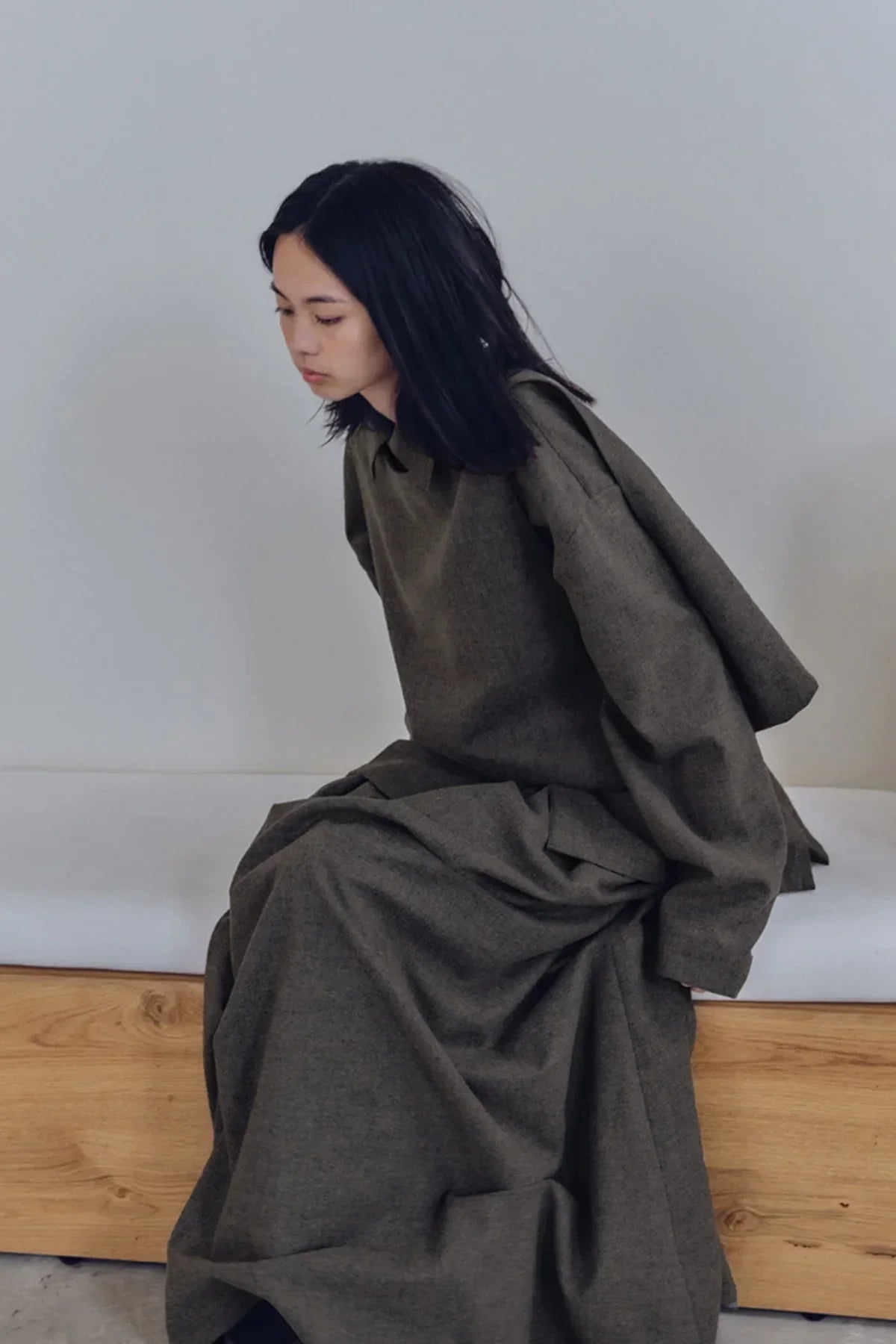 Woman with black hair wearing oversized gray outfit sitting on wooden bench with white cushion