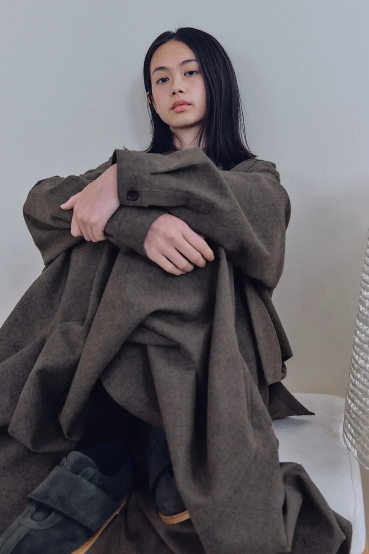 Young woman wearing oversized brown wool coat and dark shoes, sitting against plain gray wall