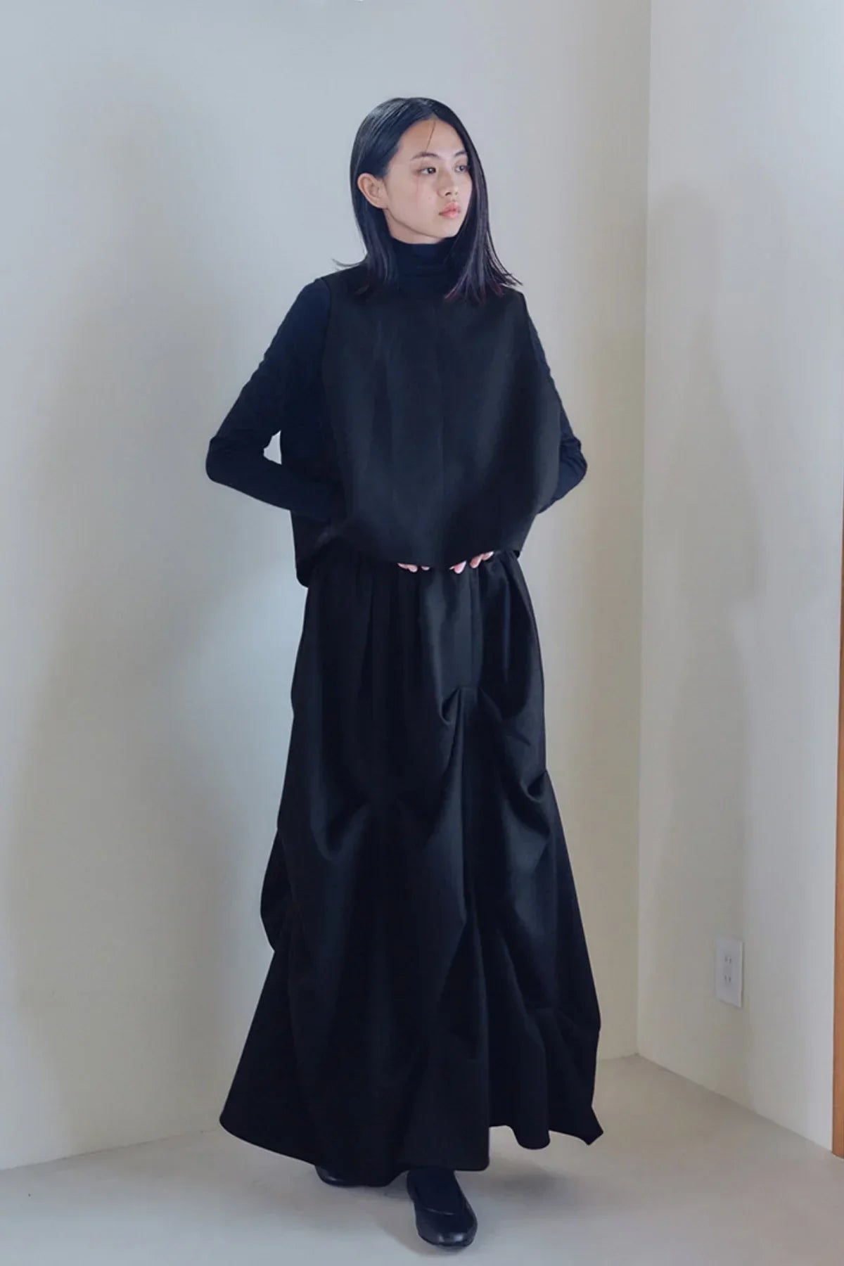 Woman wearing black turtleneck top and long black voluminous skirt standing indoors