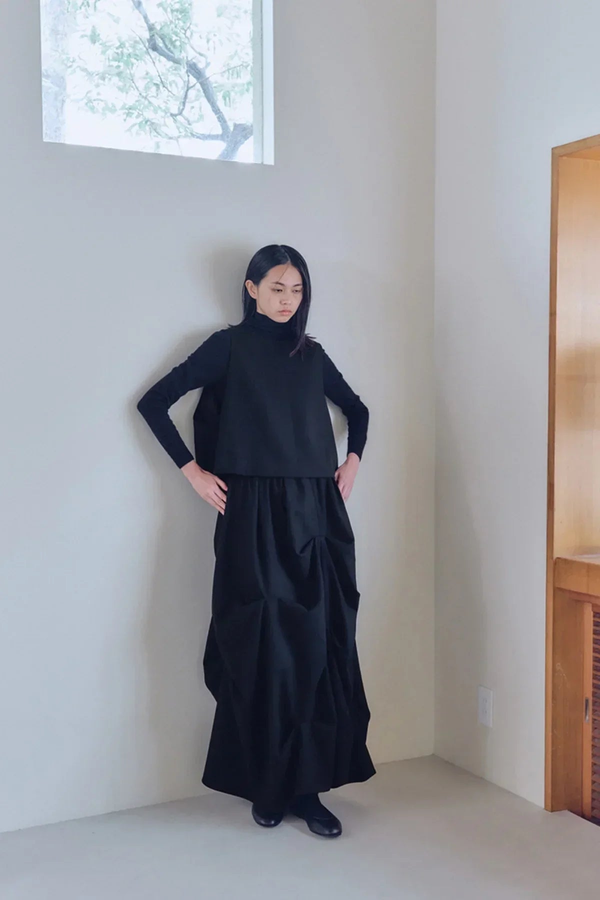 Woman wearing black layered turtleneck top and gathered long skirt in minimalist room with window