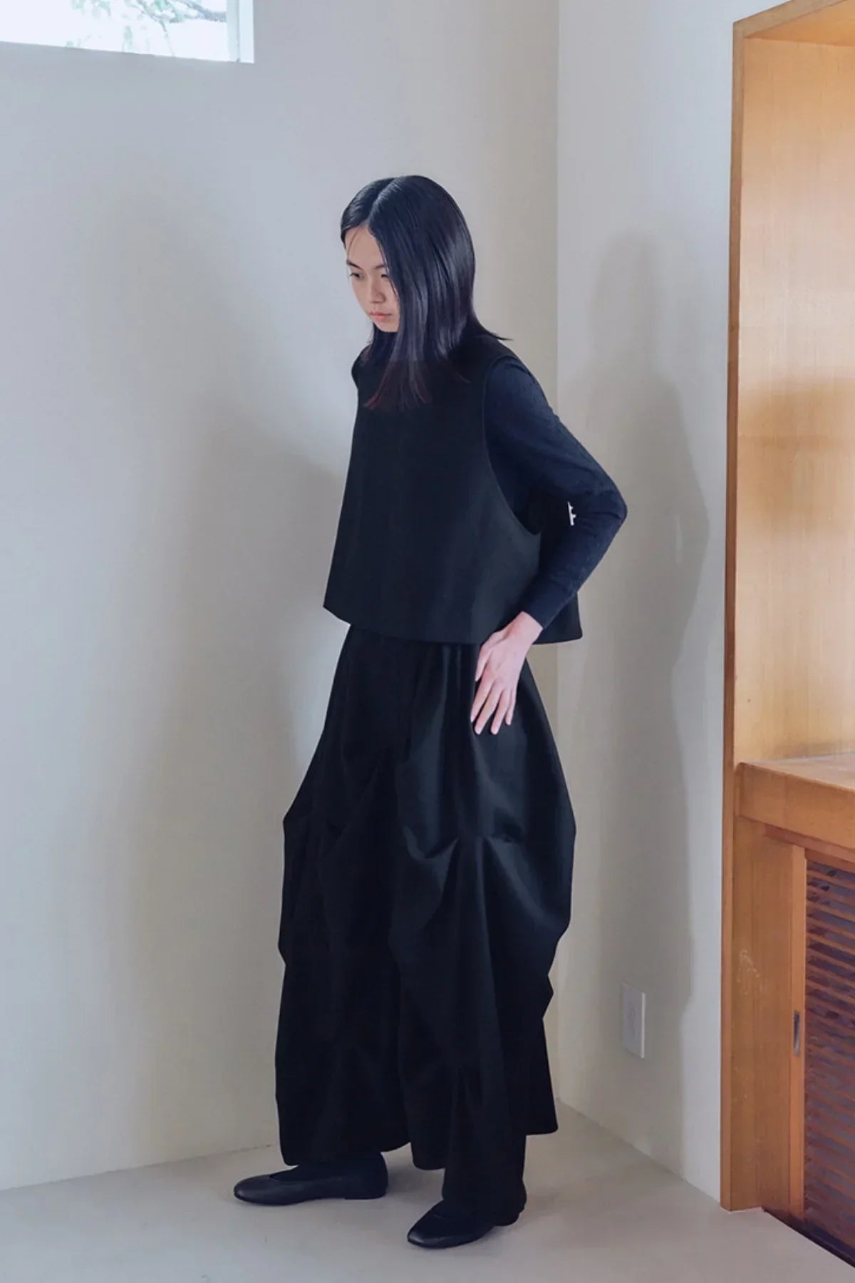 Woman wearing black sleeveless top and voluminous skirt standing indoors by wooden cabinet