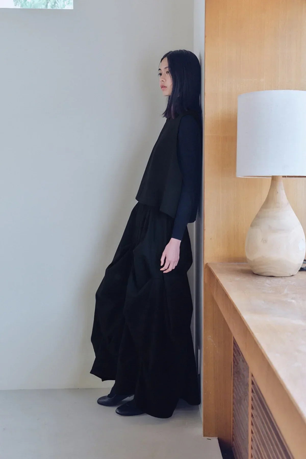 Woman wearing black sleeveless vest and voluminous black skirt leaning against a white wall indoors