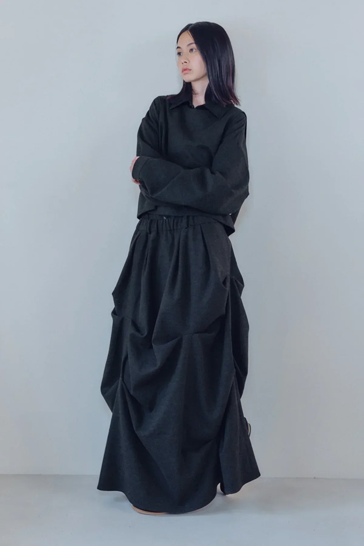 Woman wearing a charcoal oversized long skirt and matching dark long-sleeve top against plain light wall
