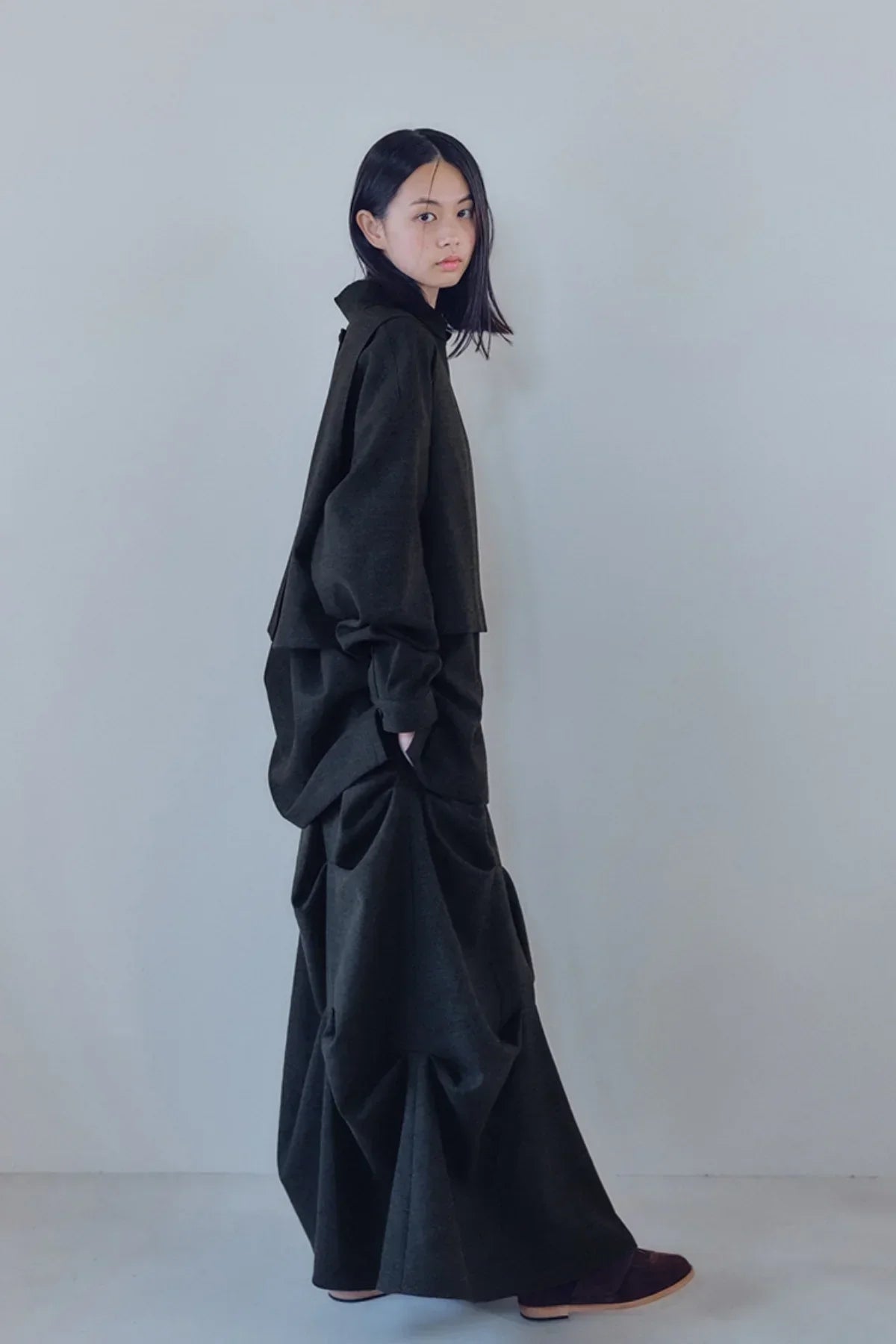 Asian woman wearing charcoal nest curtain skirt and matching oversized top against plain gray wall
