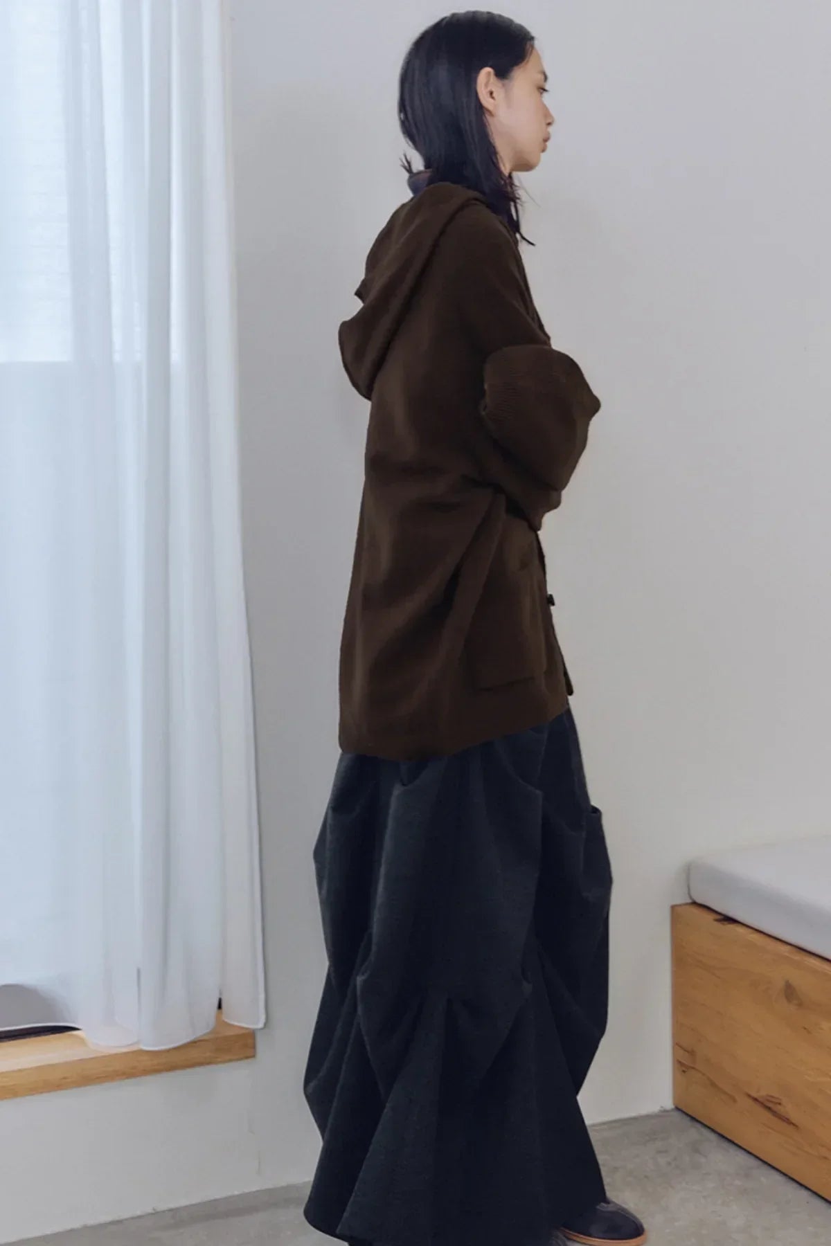 Side profile of a model wearing a dark brown hooded sweater and voluminous charcoal skirt in a minimal room