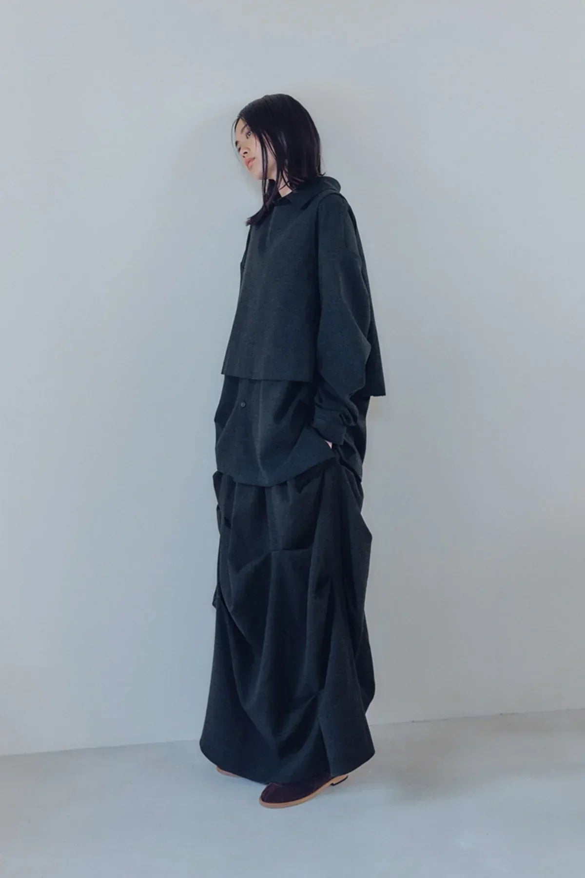 Woman wearing charcoal layered oversized vest and skirt outfit standing against a plain gray wall