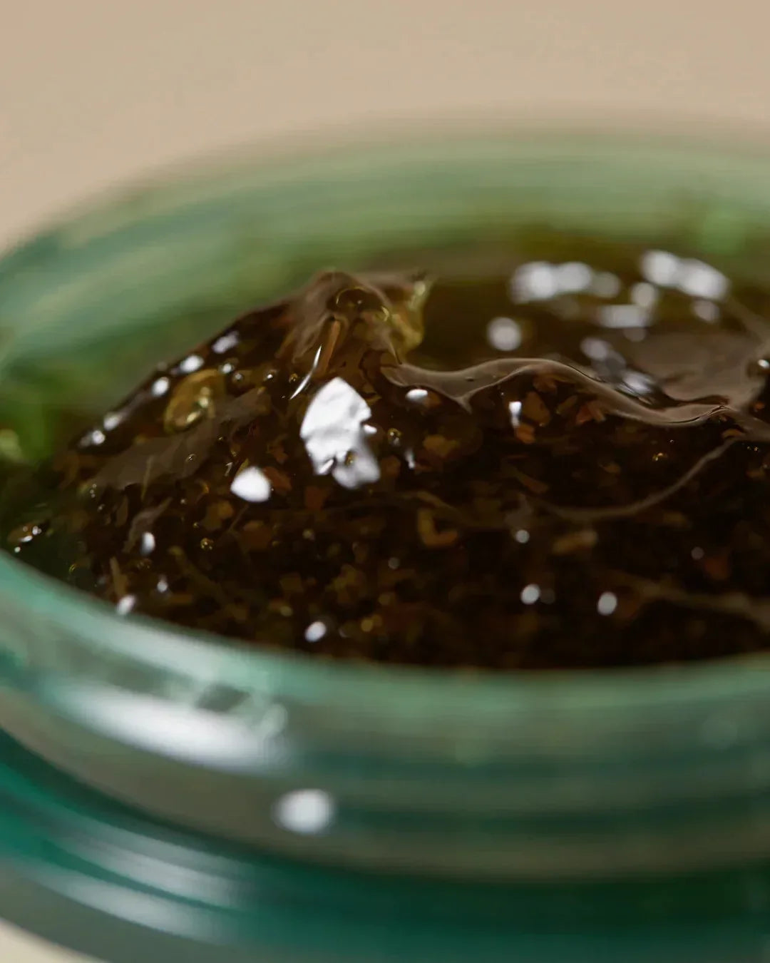 Close-up of dark gel mask with herbal particles in green glass jar on beige background