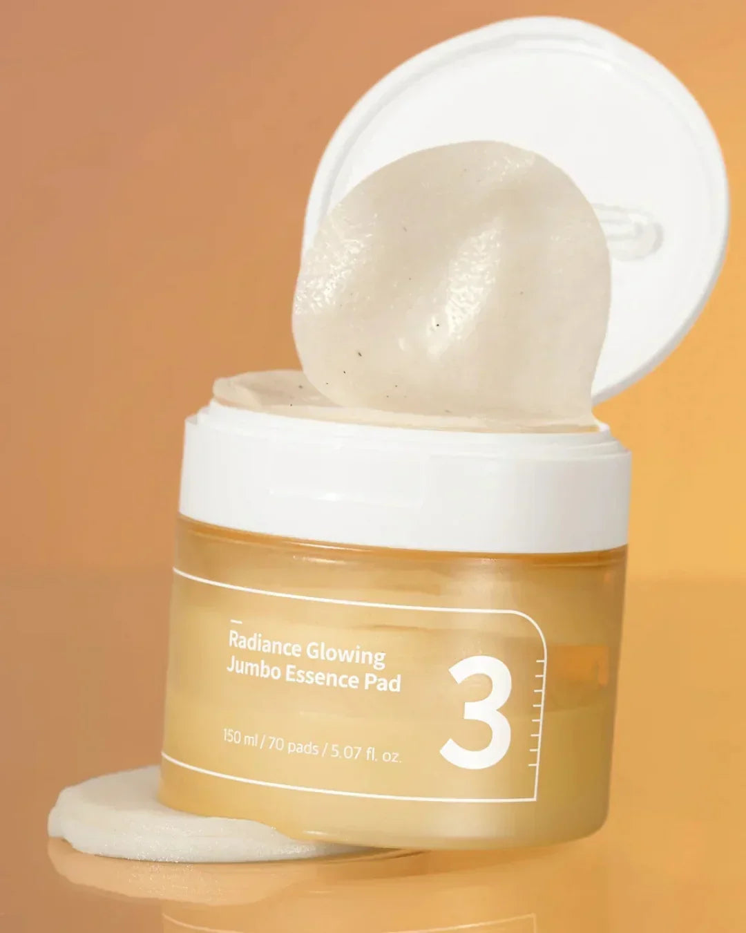 Open jar of Radiance Glowing Jumbo Essence Pad with one pad lifted, amber liquid and beige background