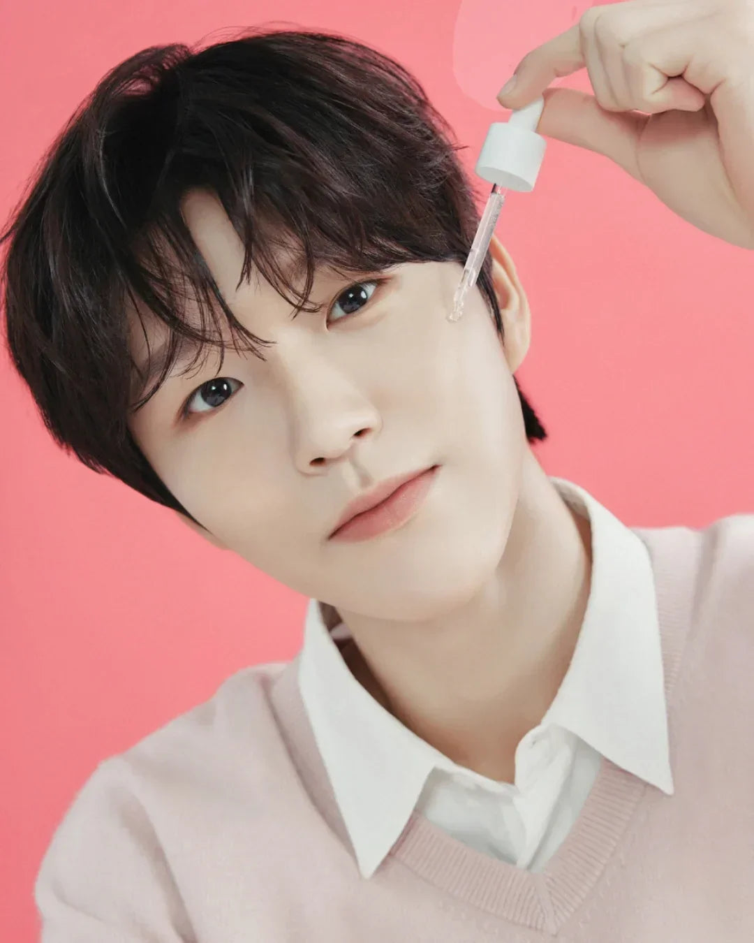 Young man applying skin softening serum with dropper on cheek against pink background