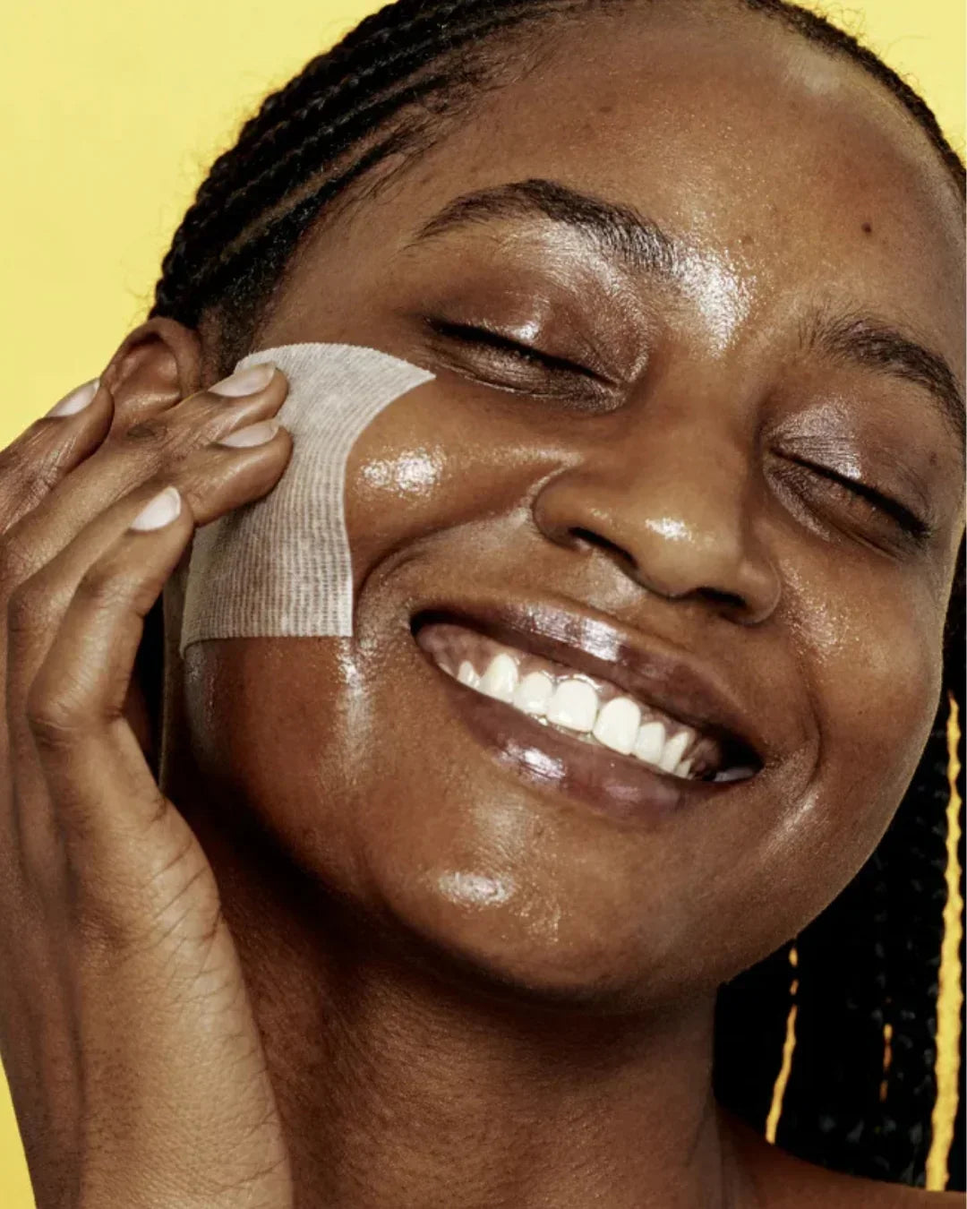 Close-up of smiling woman with glowing skin applying niacinamide pad on cheek against yellow background