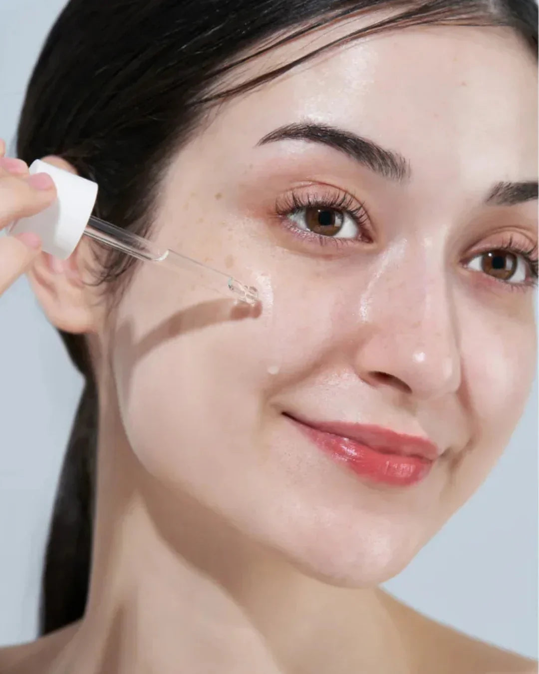 Close-up of woman applying clear facial serum with dropper to her cheek, glowing skin