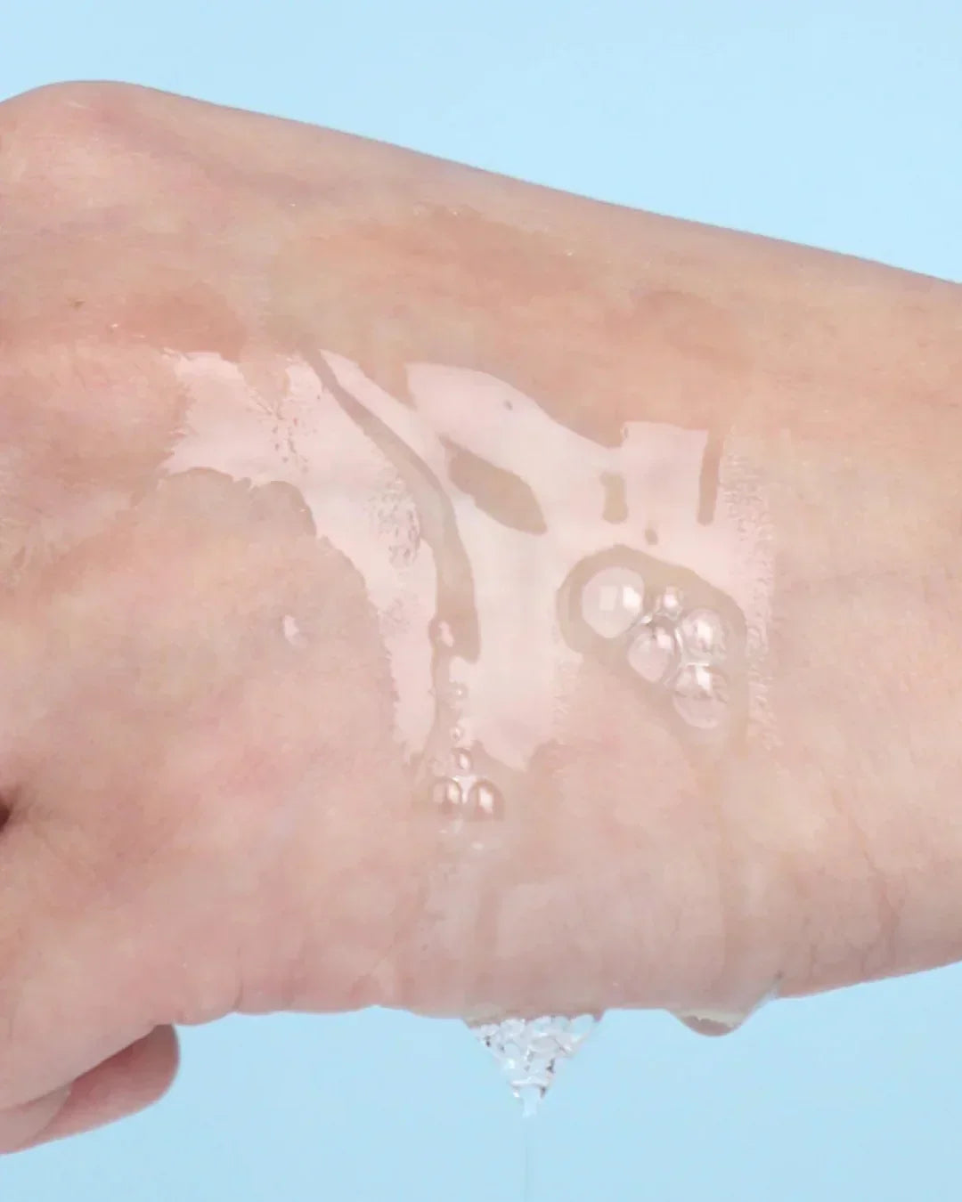 Clear serum texture on hand over light blue background showing hydrating skincare essence