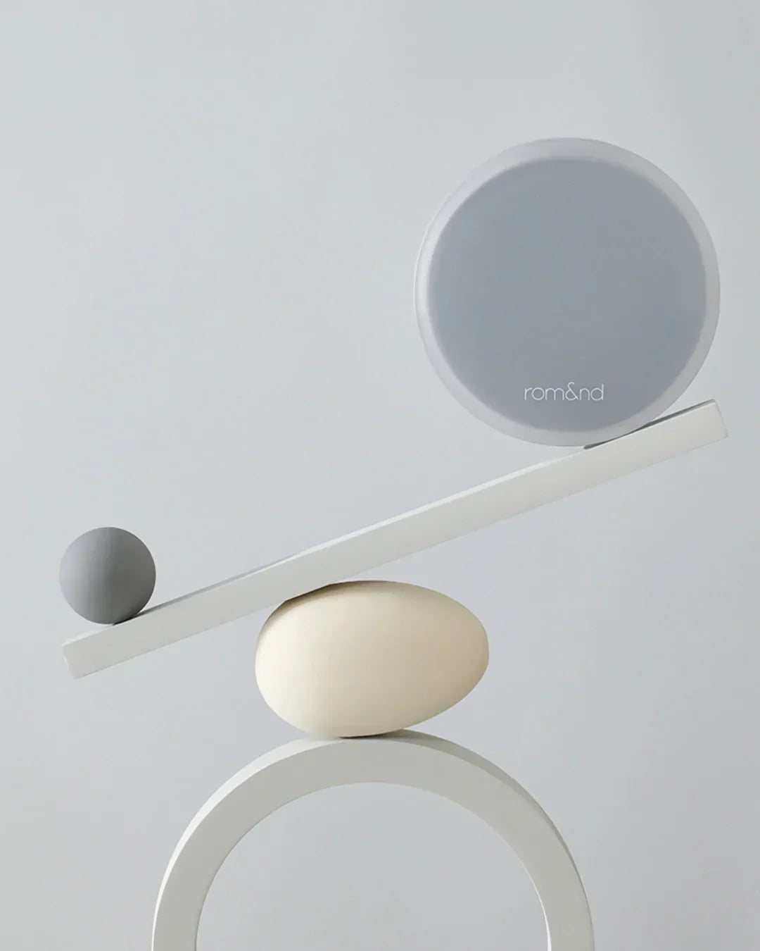 Minimalist studio composition with grey round Rom&nd case and small grey ball balanced on white shapes