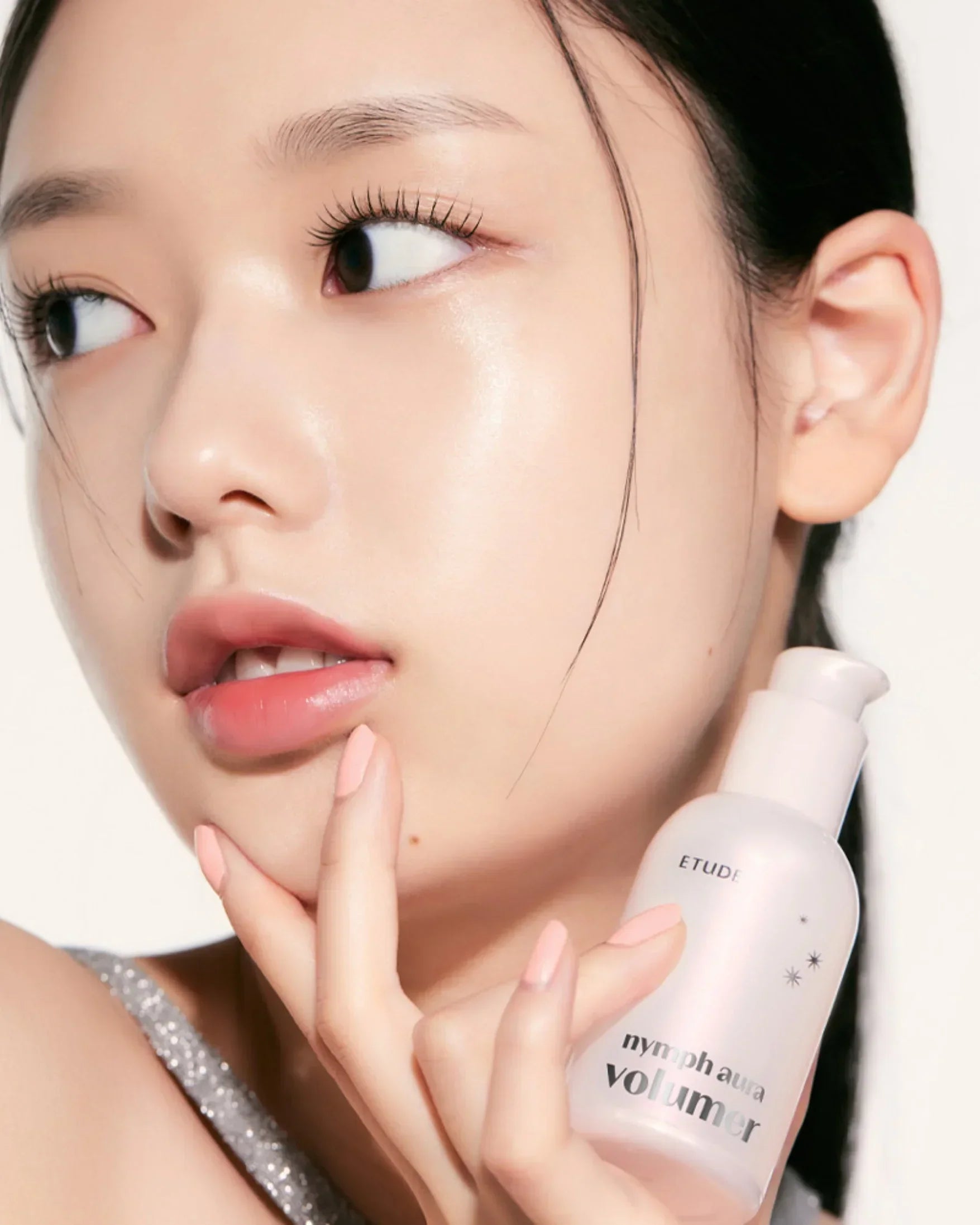 Close-up of woman with glowing skin holding Etude Nymph Aura Volumer skincare bottle