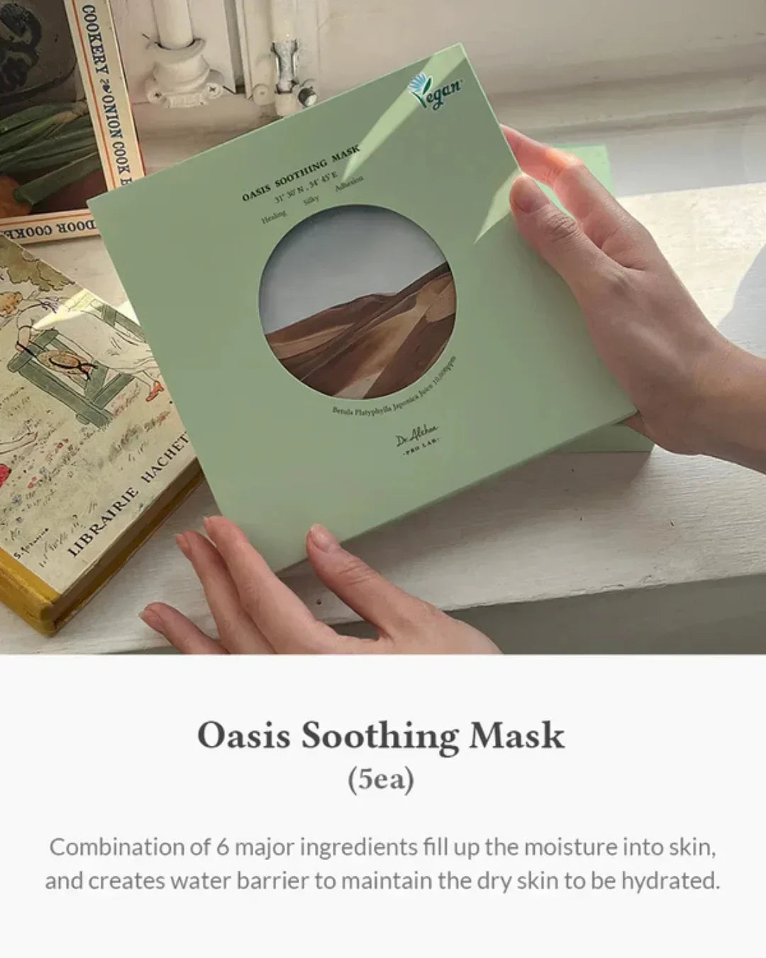 Hands holding a green box of vegan Oasis Soothing Mask skincare with desert illustration on window sill