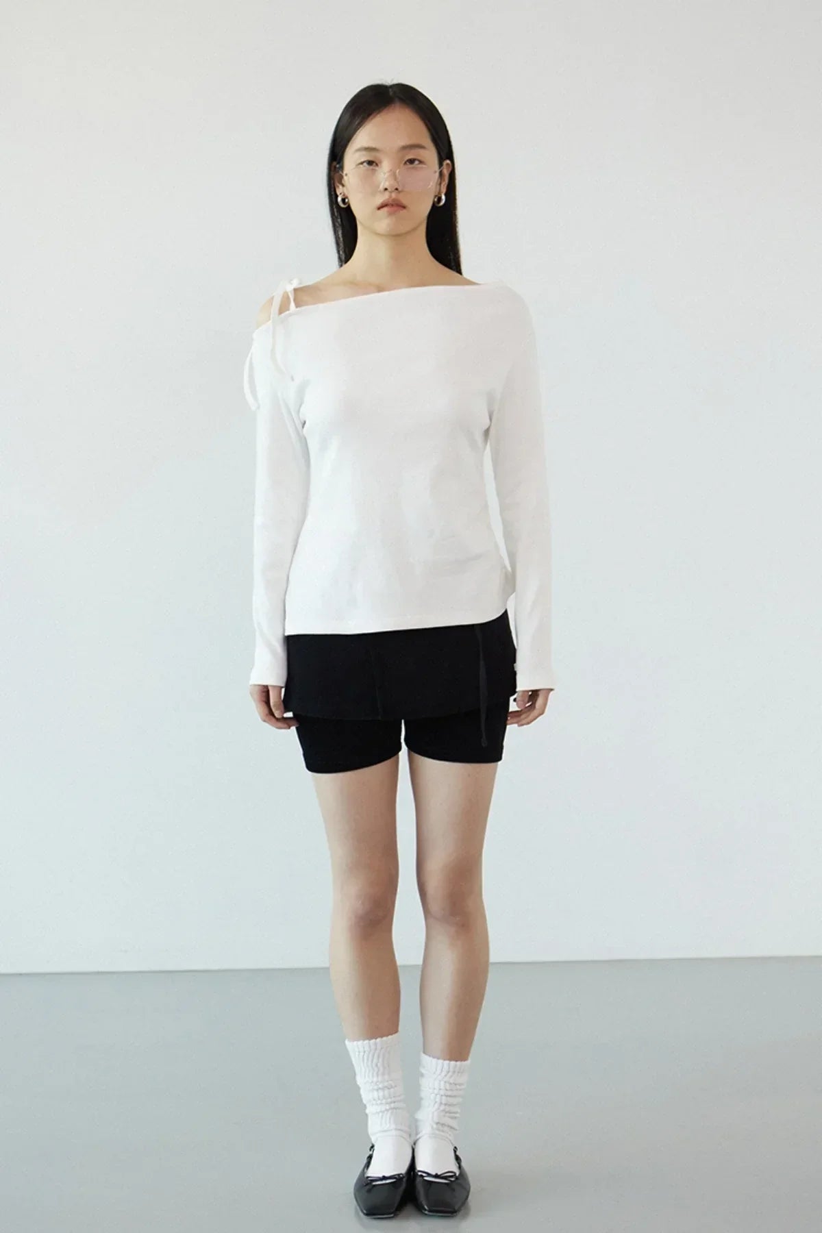 Model wearing off-shoulder ivory long sleeve top with black shorts, white socks, and black flats