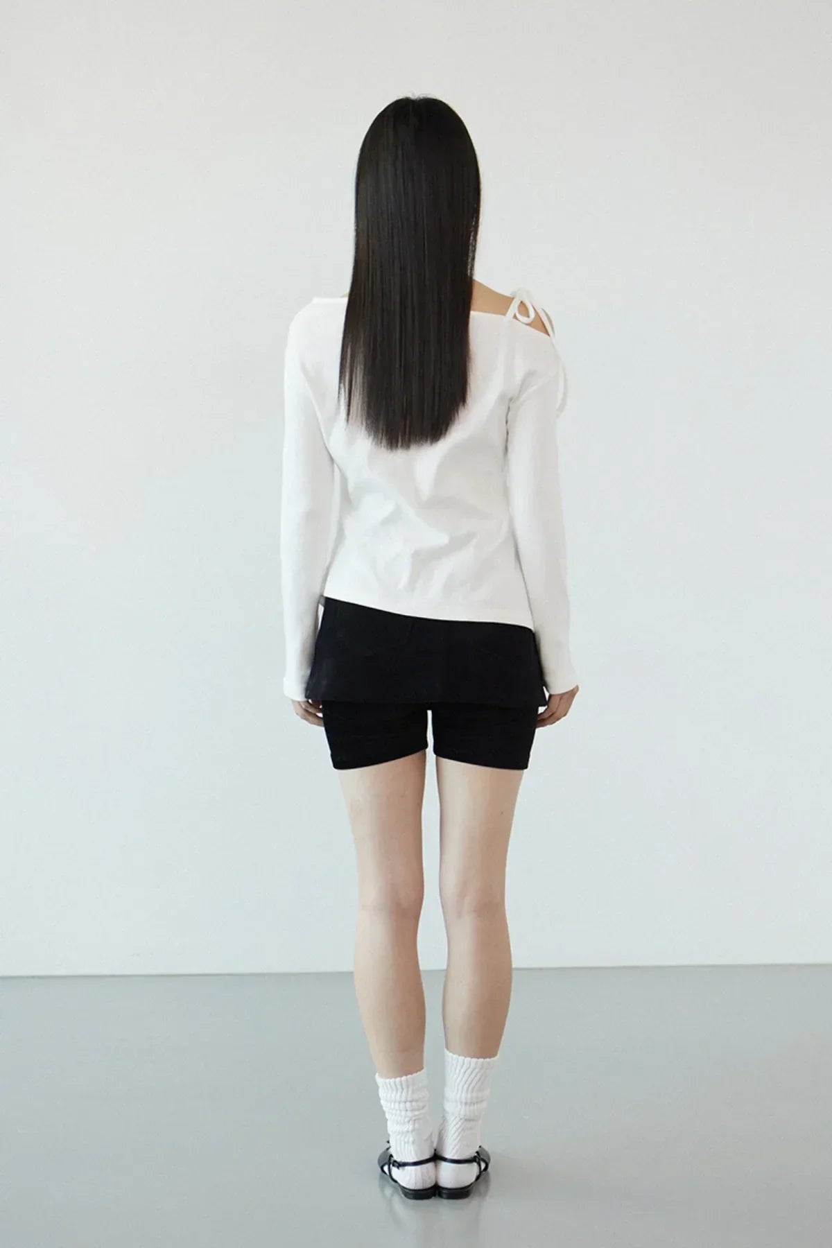 Woman standing facing away in white off-shoulder long sleeve top, black shorts, white socks, and black sandals