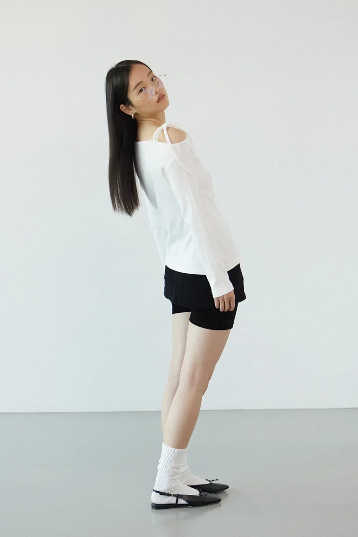 Woman in off-shoulder white long-sleeve top, black shorts, white socks, and black flats against plain background