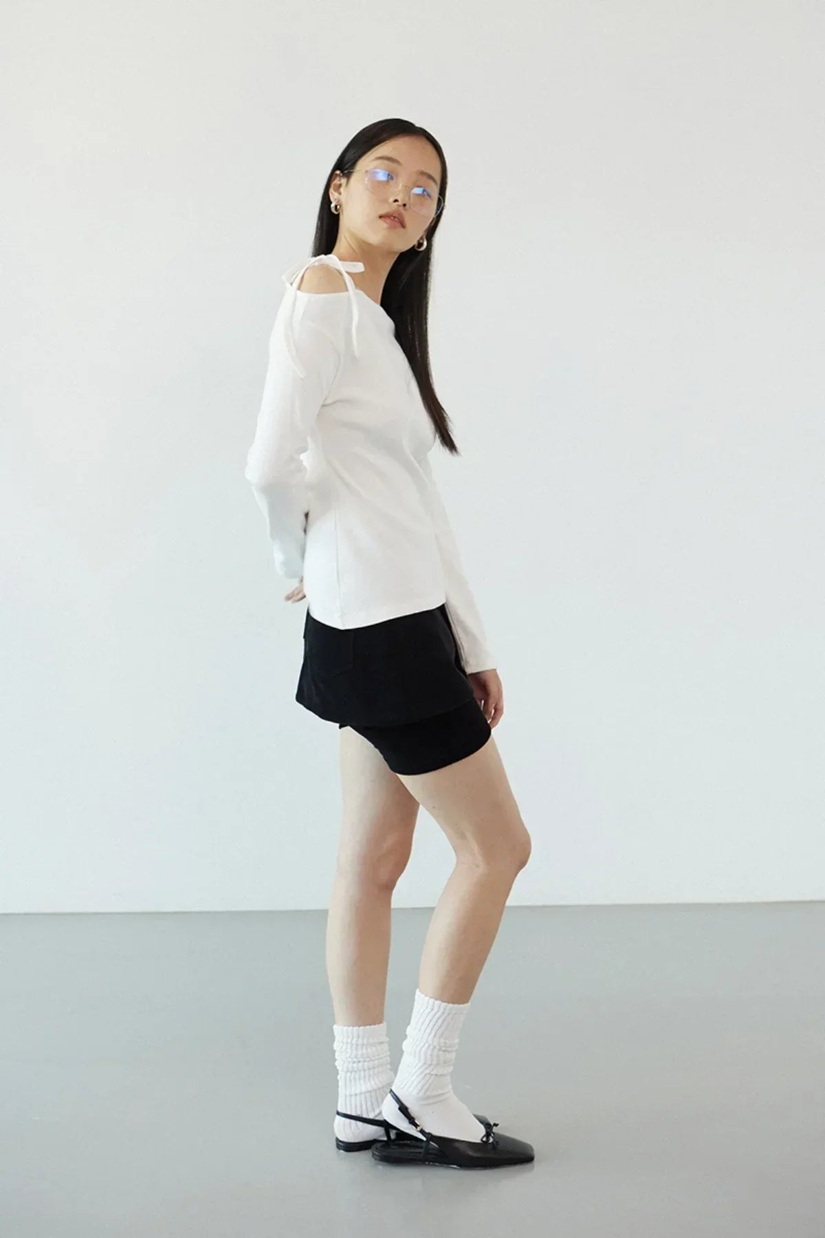 Woman wearing ivory off-shoulder long sleeve top, black mini skirt, white socks, and black flats in studio