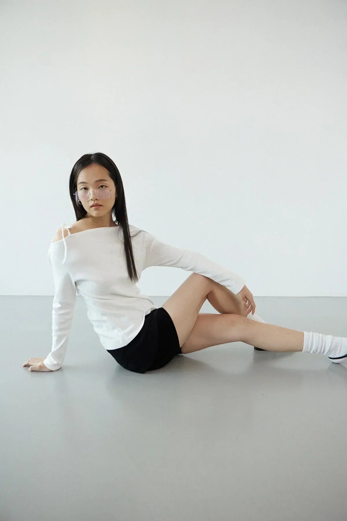 Young woman in off-shoulder white long-sleeve top and black shorts sitting on gray floor in minimal studio