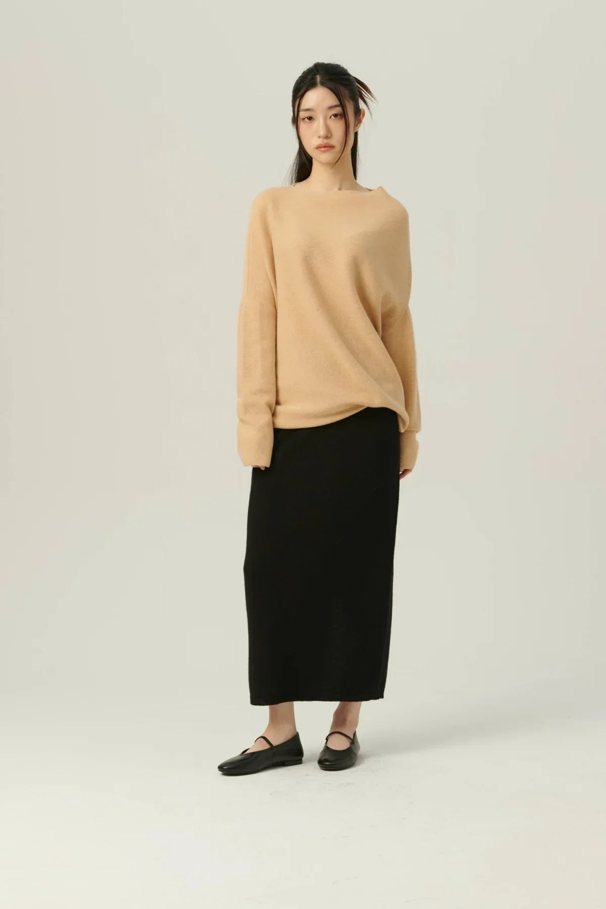 Woman in beige oversized sweater, black midi skirt, and black flats on neutral background